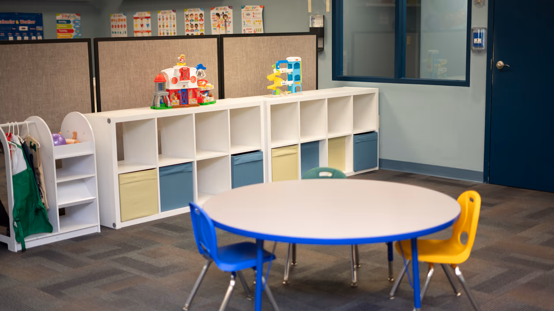 Indoor activity room with round table, blue and yellow chairs, and cubby storage.