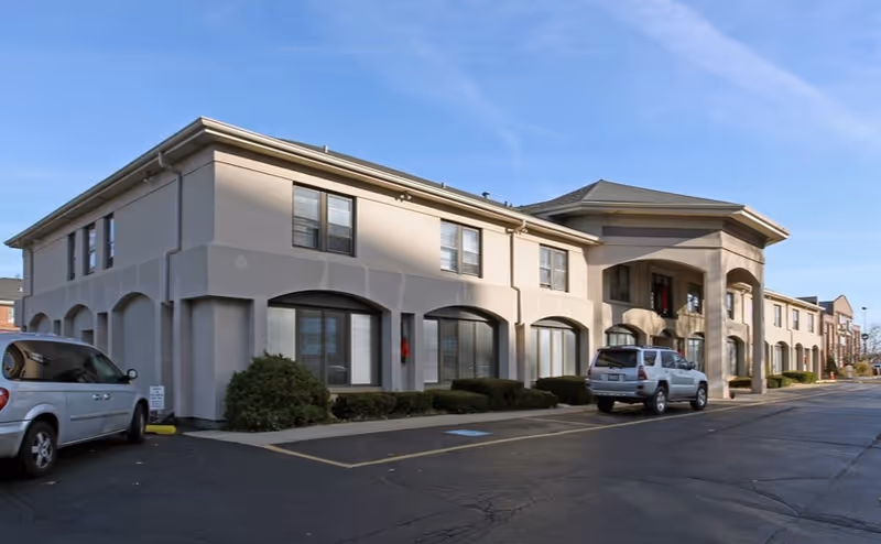 Two-story beige senior living building with arched windows and parked cars along the driveway.