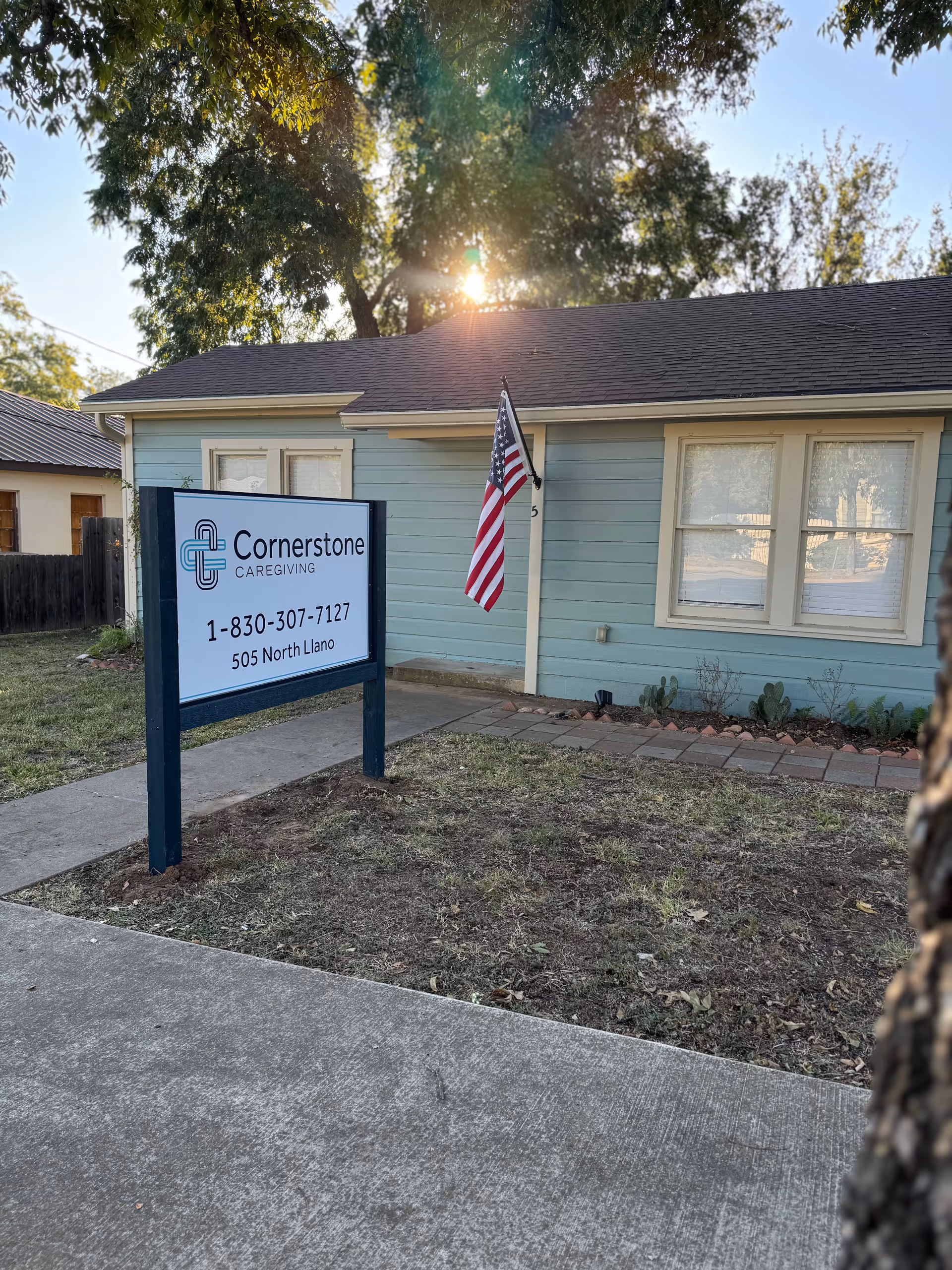 Light blue, single-story facility exterior with a Cornerstone Caregiving sign and an American flag.