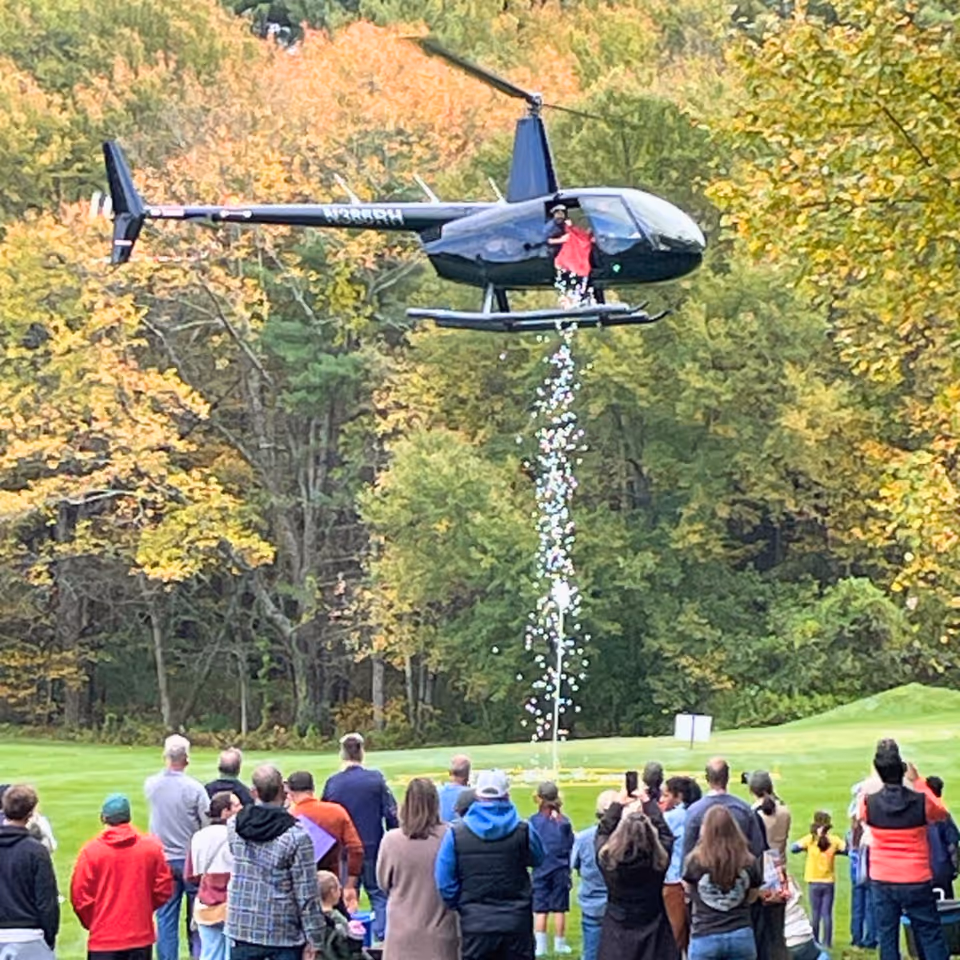 A dark helicopter hovers above a crowd on a grassy field, releasing a stream downward.