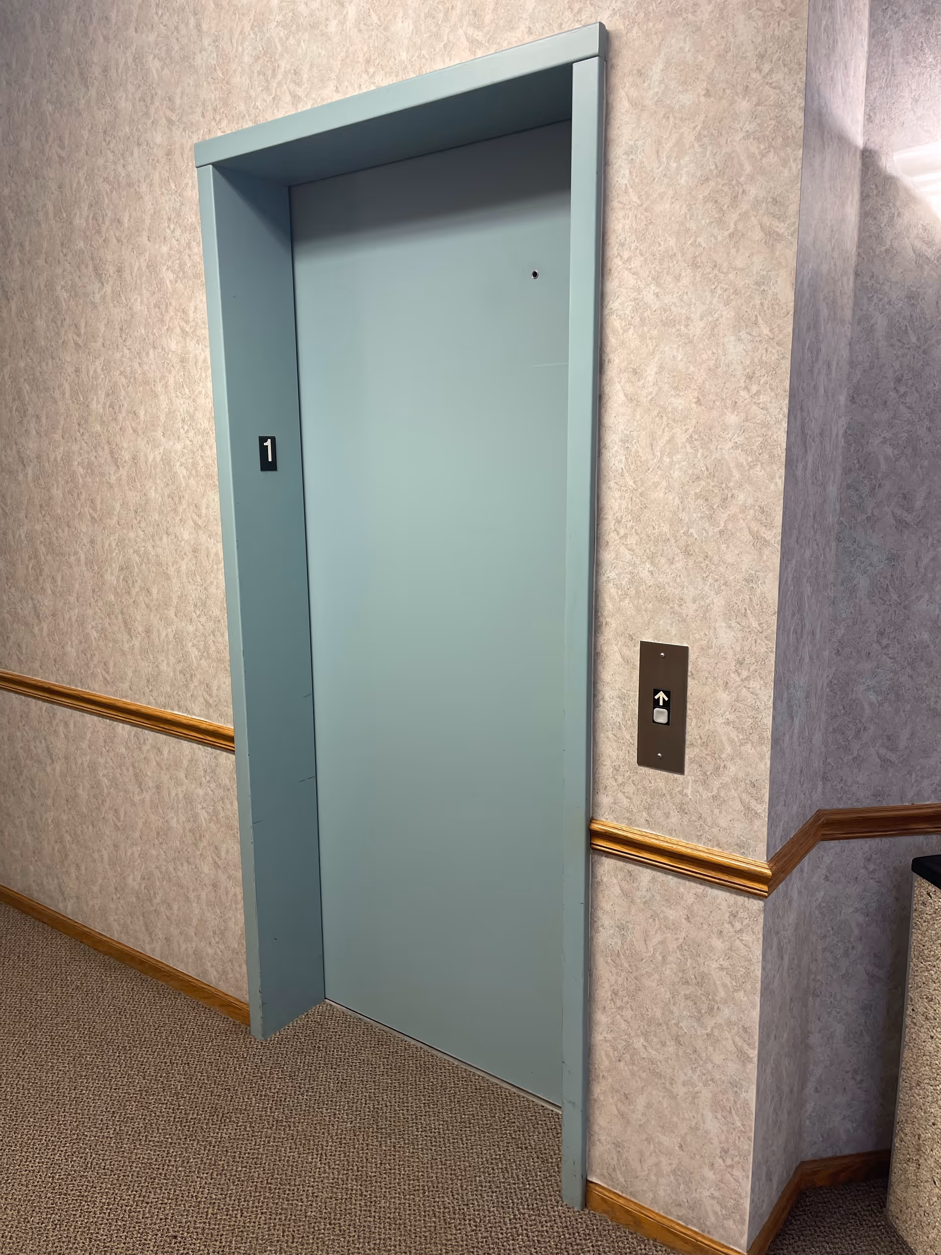 A blue elevator door sits in a hallway with textured walls and wood trim.