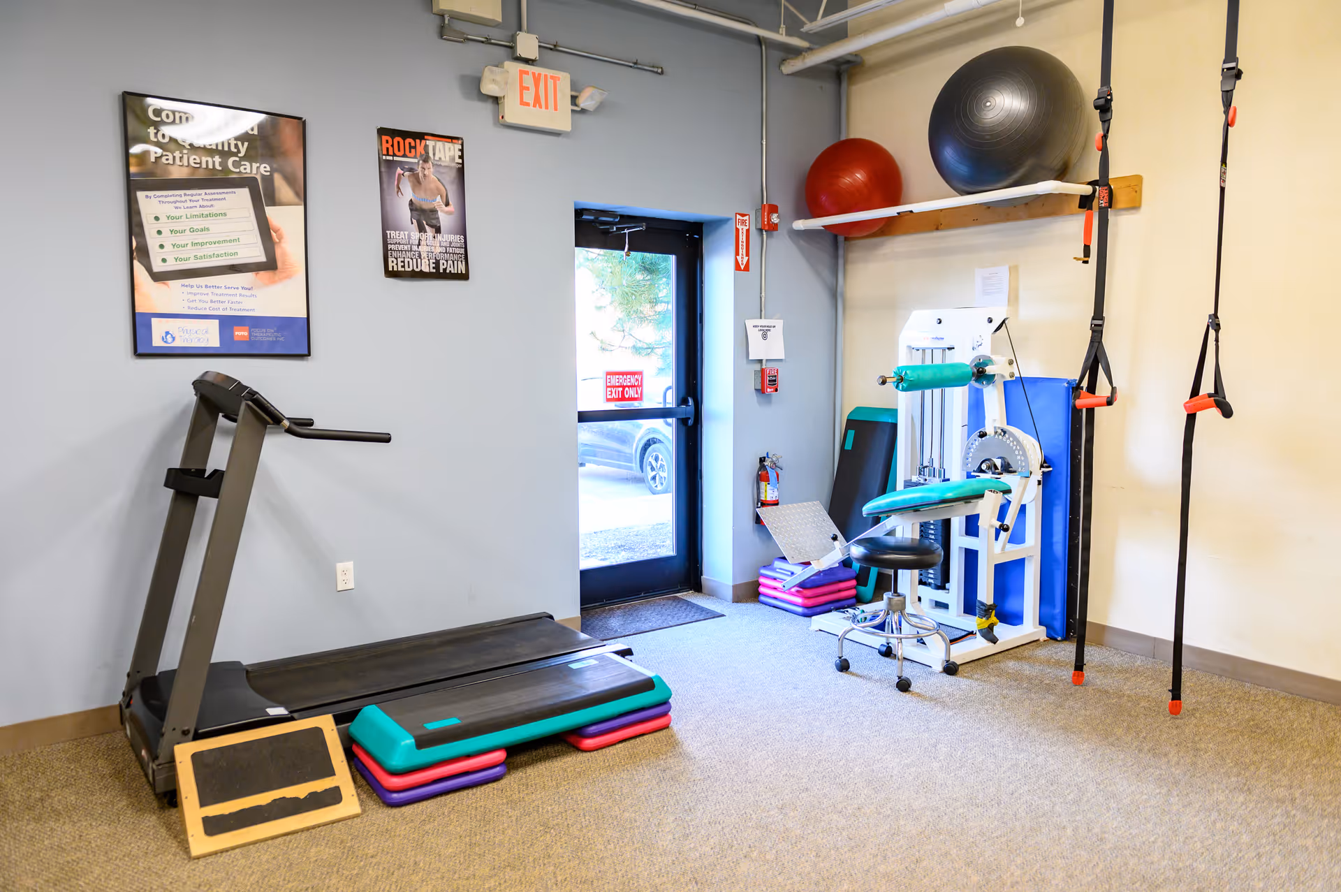Small physical therapy gym with treadmill, exercise machines, mats, resistance bands, TRX straps, and a door.