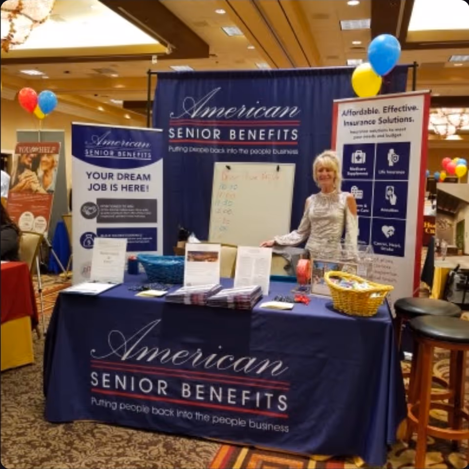 A smiling woman stands beside an American Senior Benefits booth at an indoor expo.