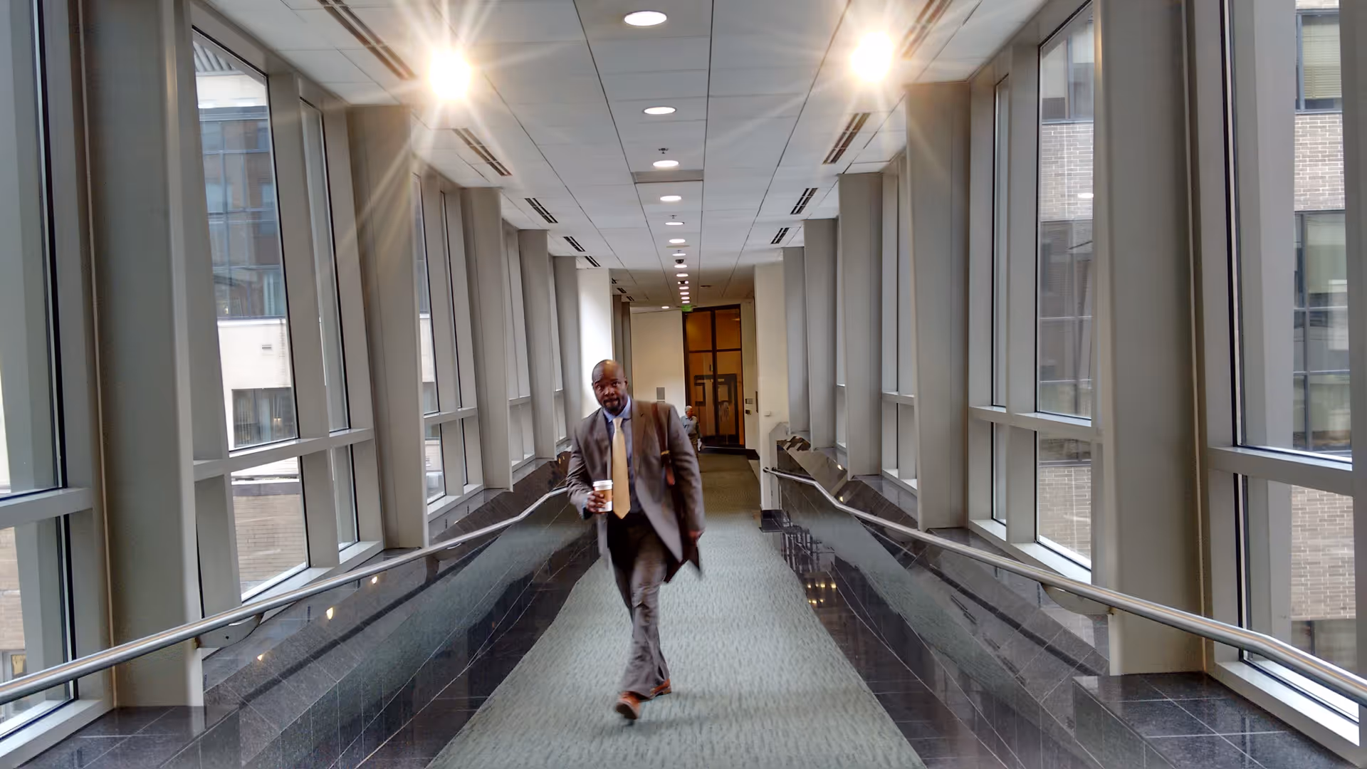 A man in a suit walks down a bright, window-lined hospital hallway, coffee cup in hand.