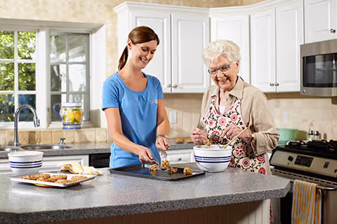 A caregiver and elderly woman cook together in a bright kitchen.