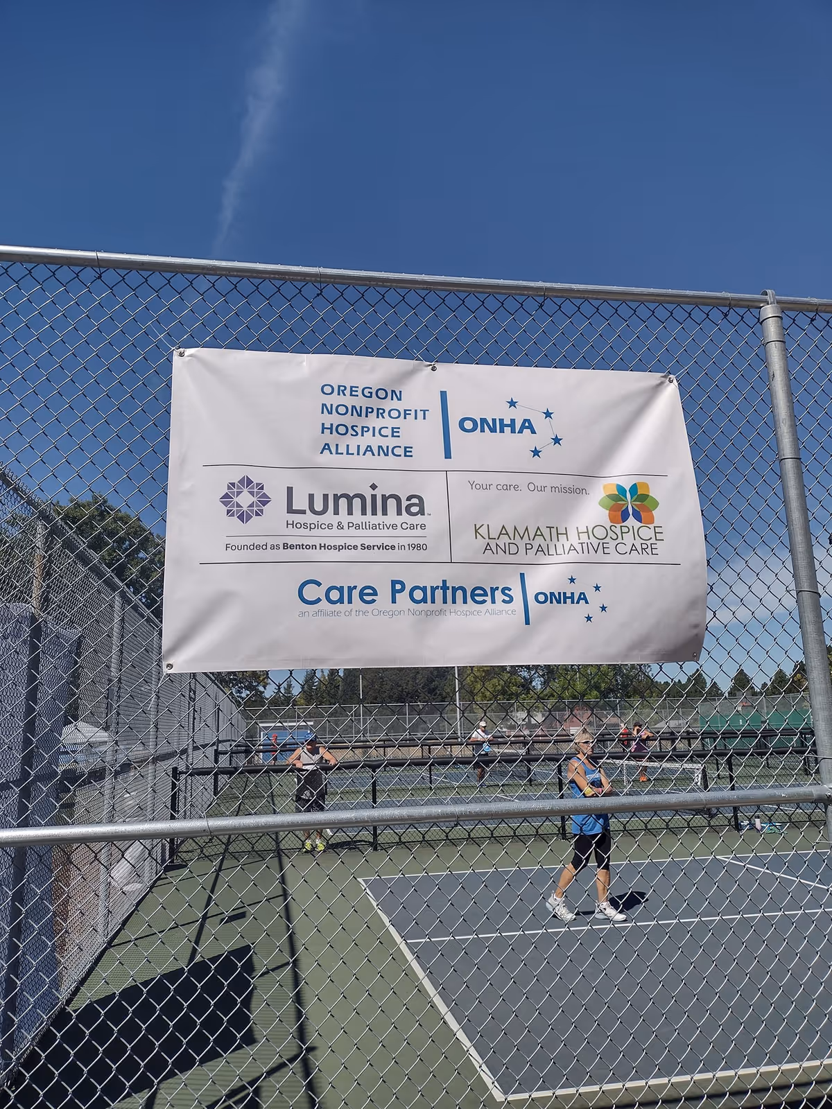 Chain-link fence with a Lumina Hospice banner over an outdoor tennis court.