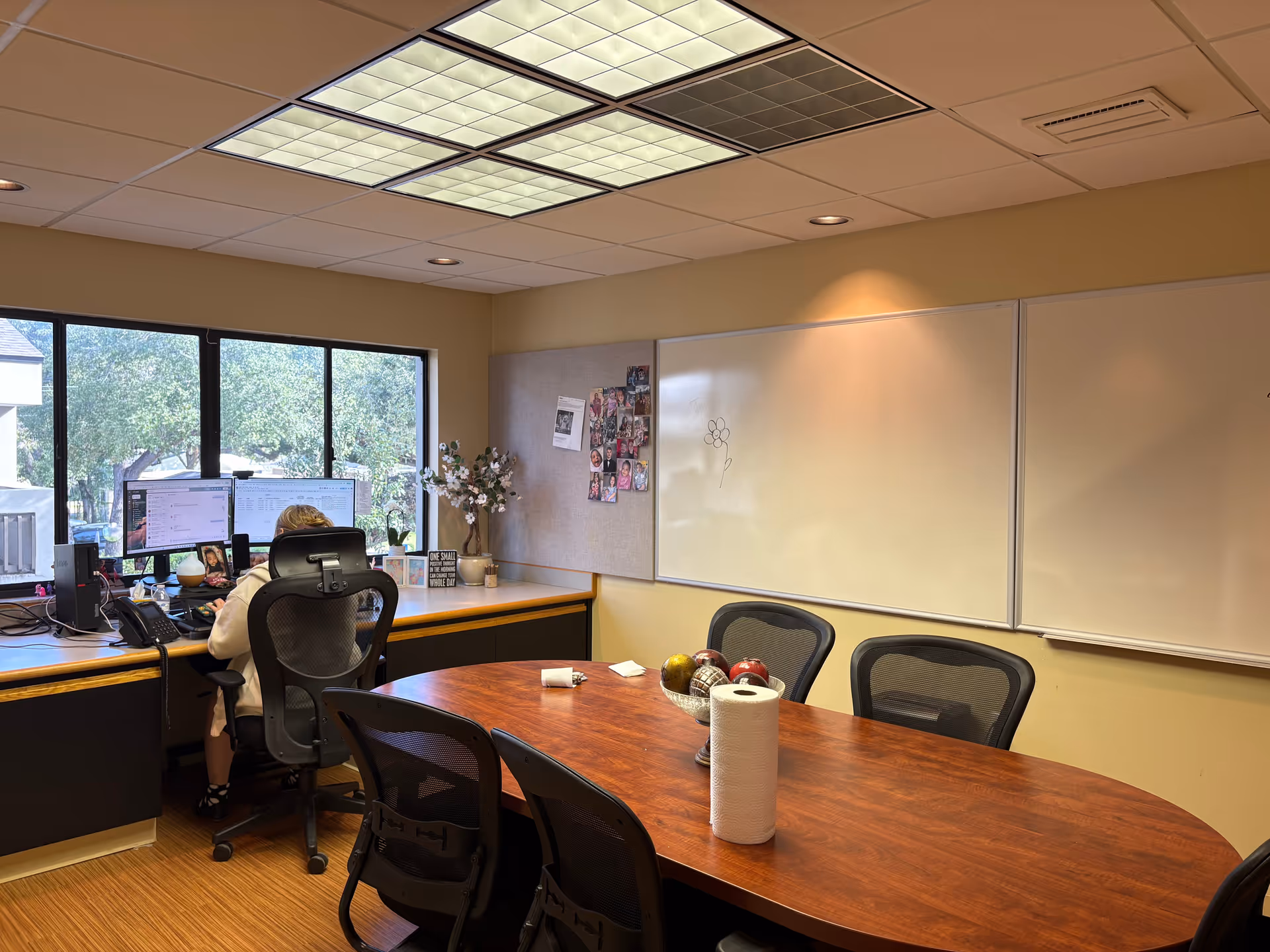 Office meeting room with a long wooden table, whiteboards, and a staffer at a computer.