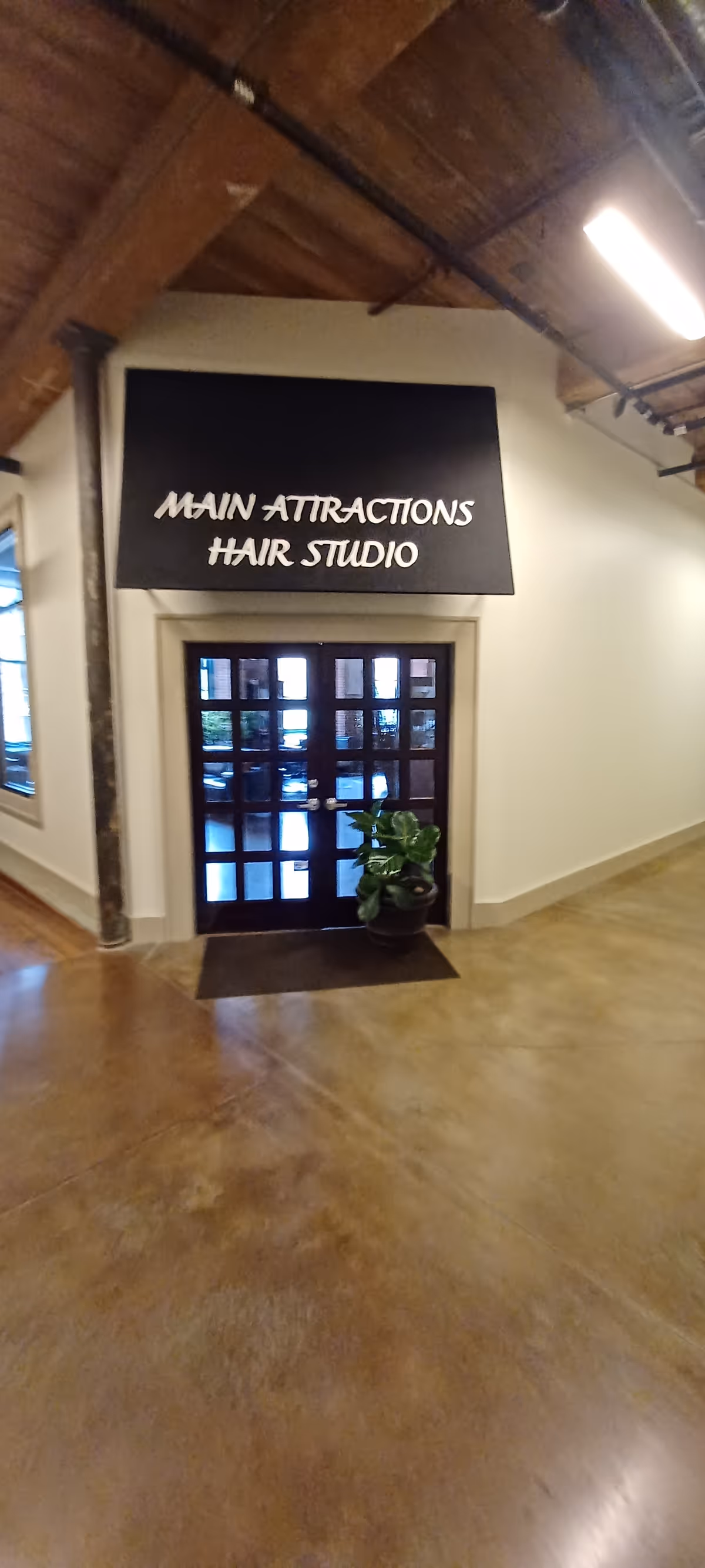 Interior hallway with a black sign Main Attractions Hair Studio above a glass door and plant.