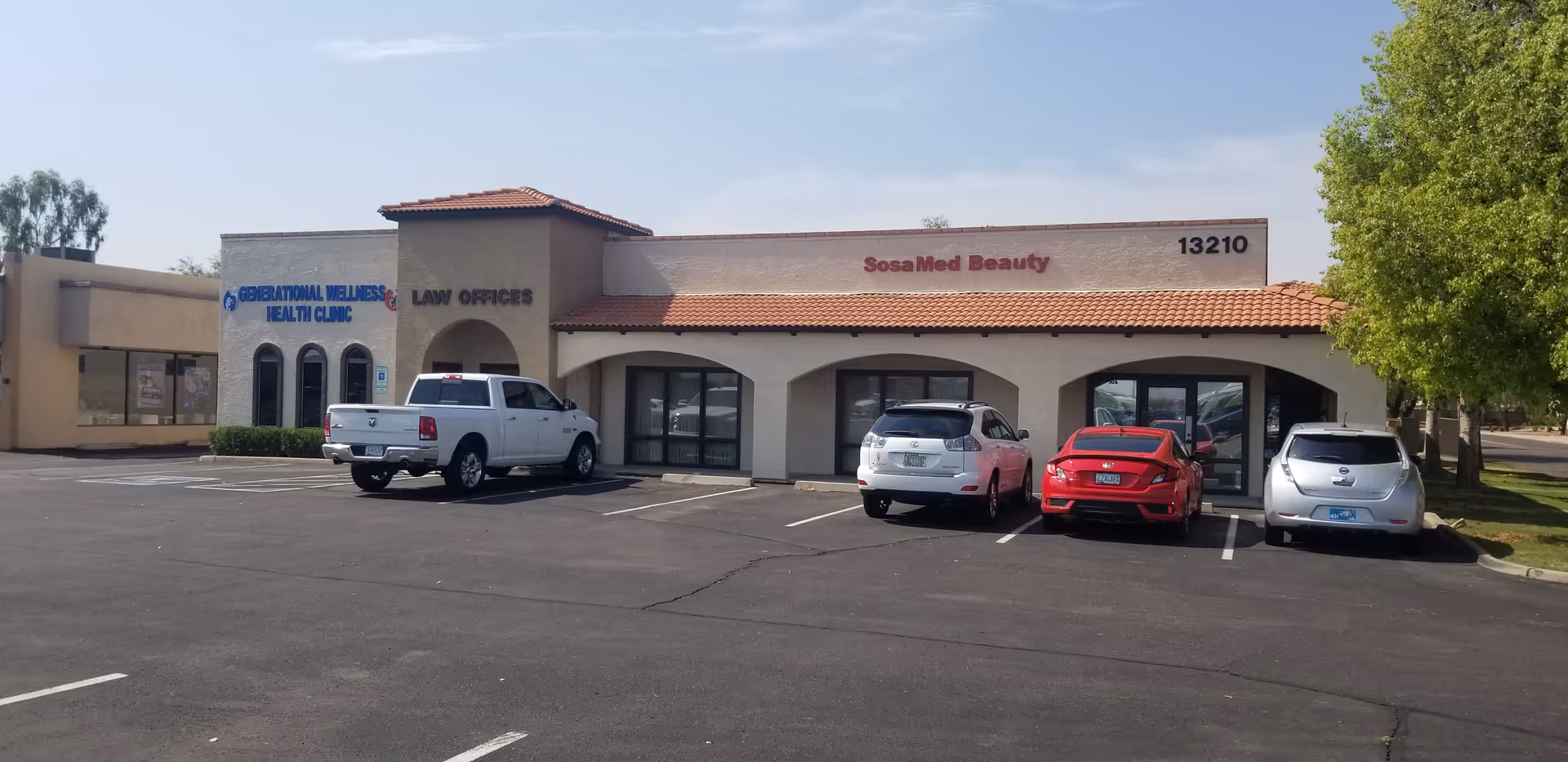 Exterior view of a beige strip plaza with arched storefronts and parked cars.