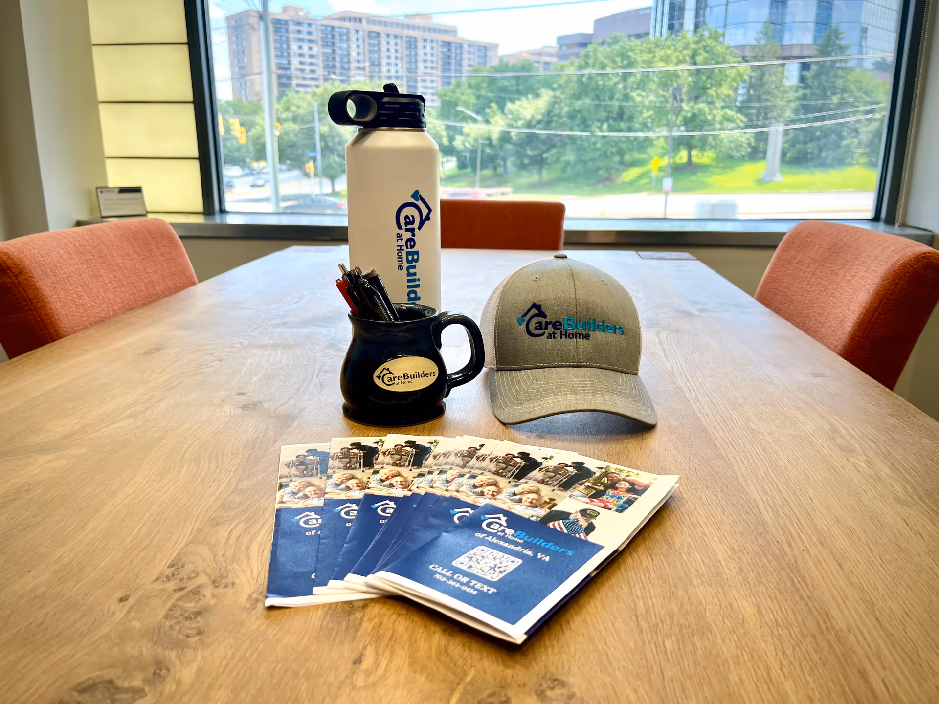 Table with CareBuilders at Home brochures, cap, mug, and water bottle by window.
