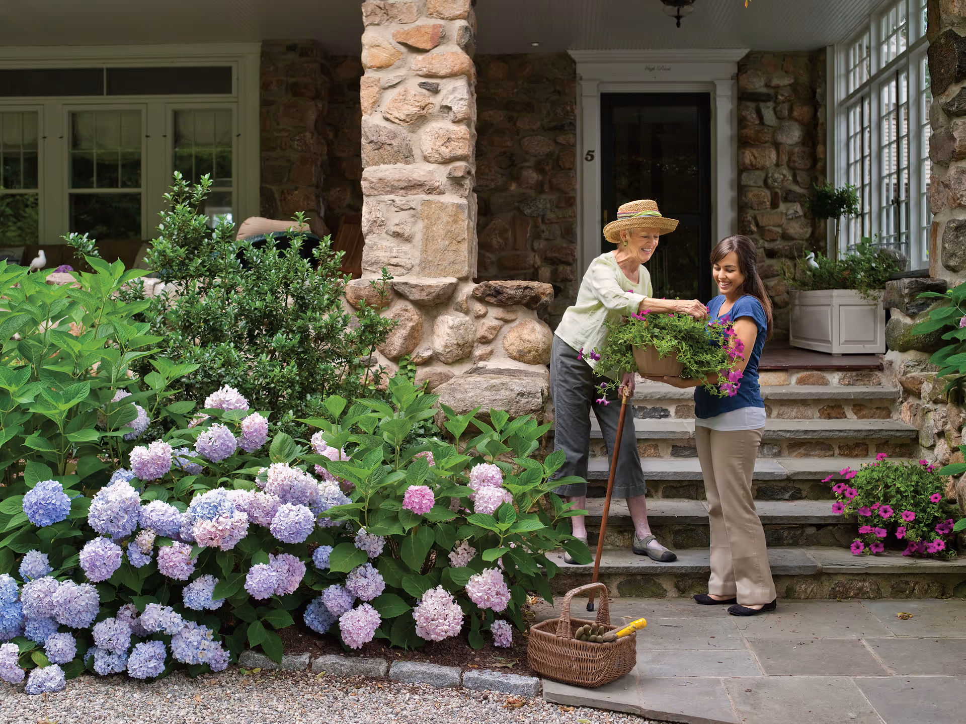Two women on a stone-front porch share a pot of flowers beside blooming hydrangeas.