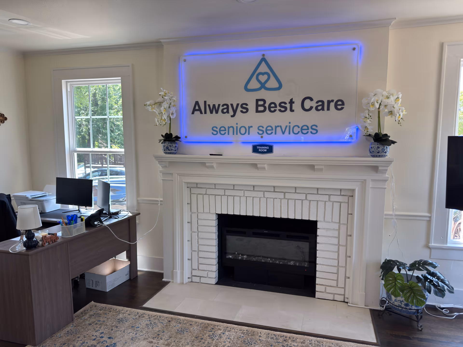 Lobby area with a fireplace mantel, blue neon Always Best Care sign, and desk.
