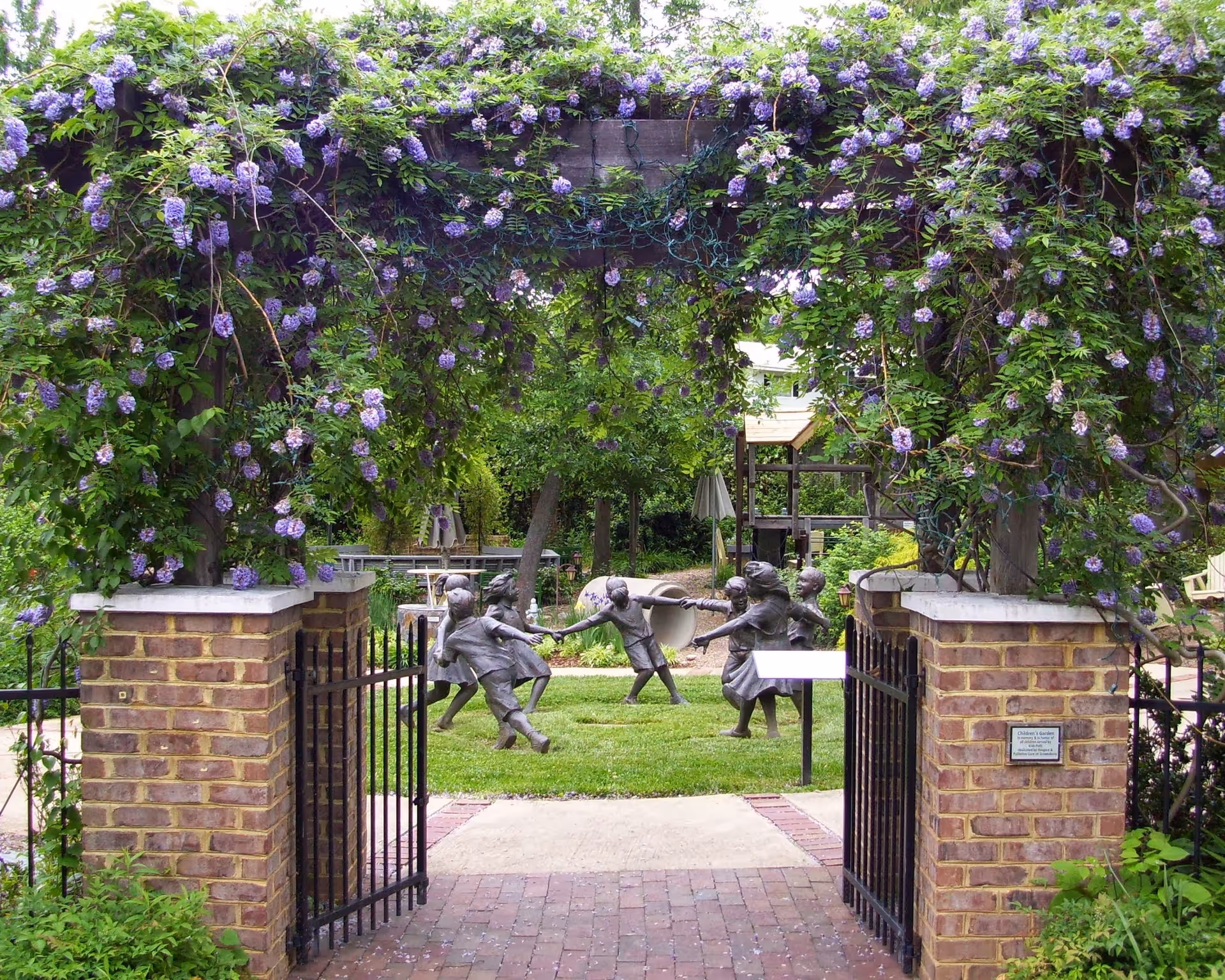 Brick arch covered with purple flowering vines frames open gate to a garden with children's statues.