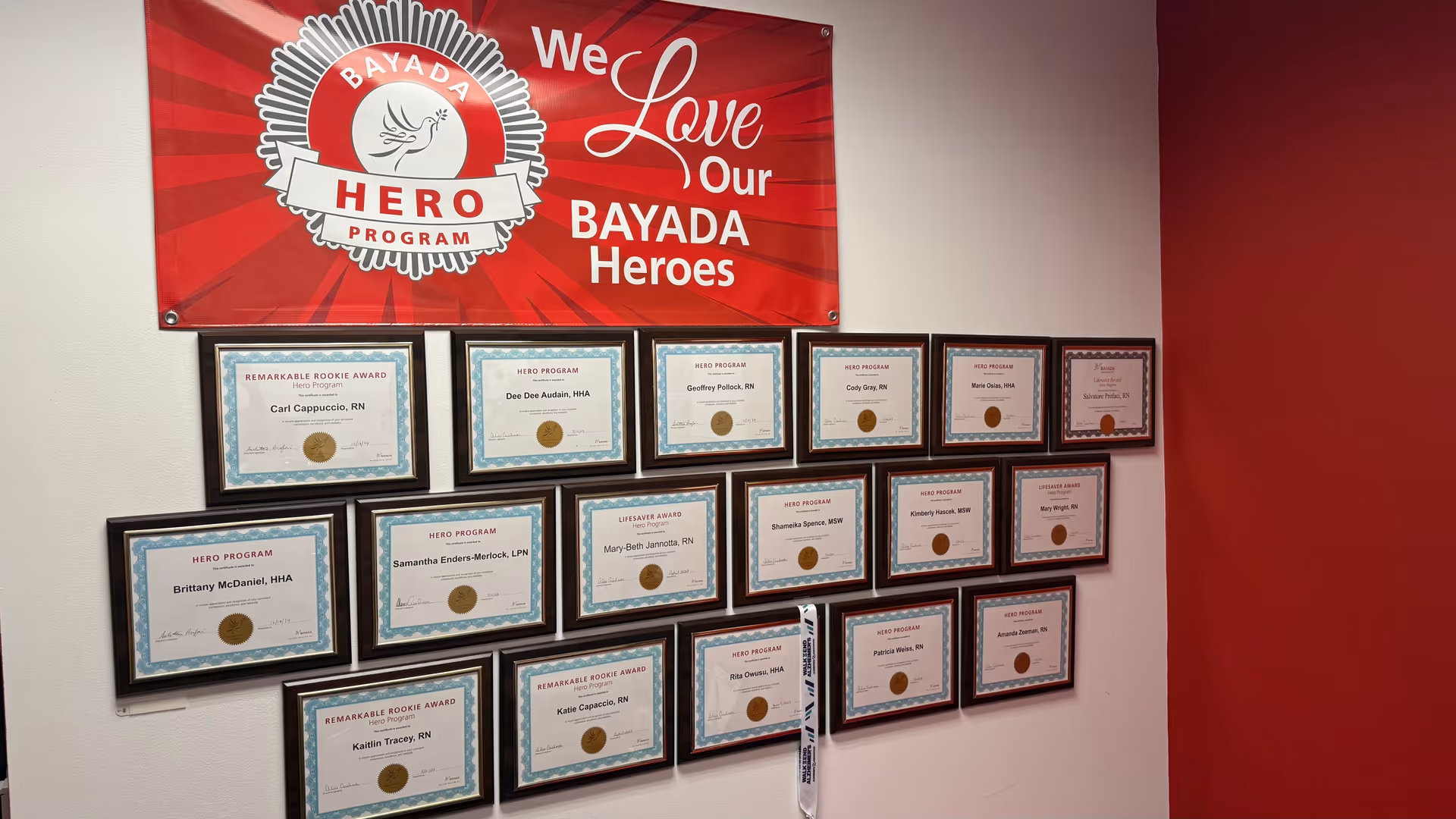 Indoor wall shows a red BAYADA banner and rows of framed certificates.
