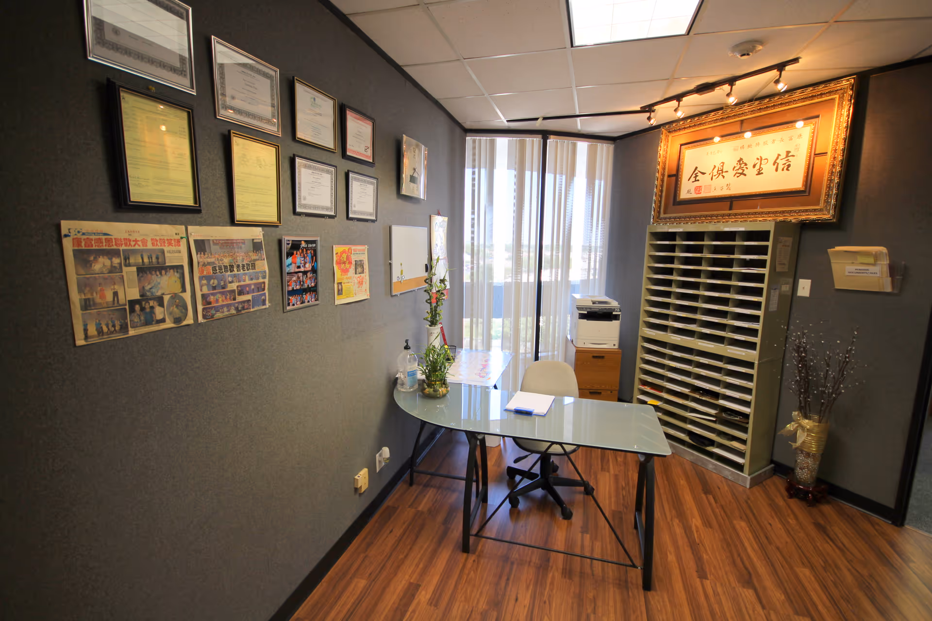Office workspace with framed certificates on a gray wall, glass desk, and file cabinets.