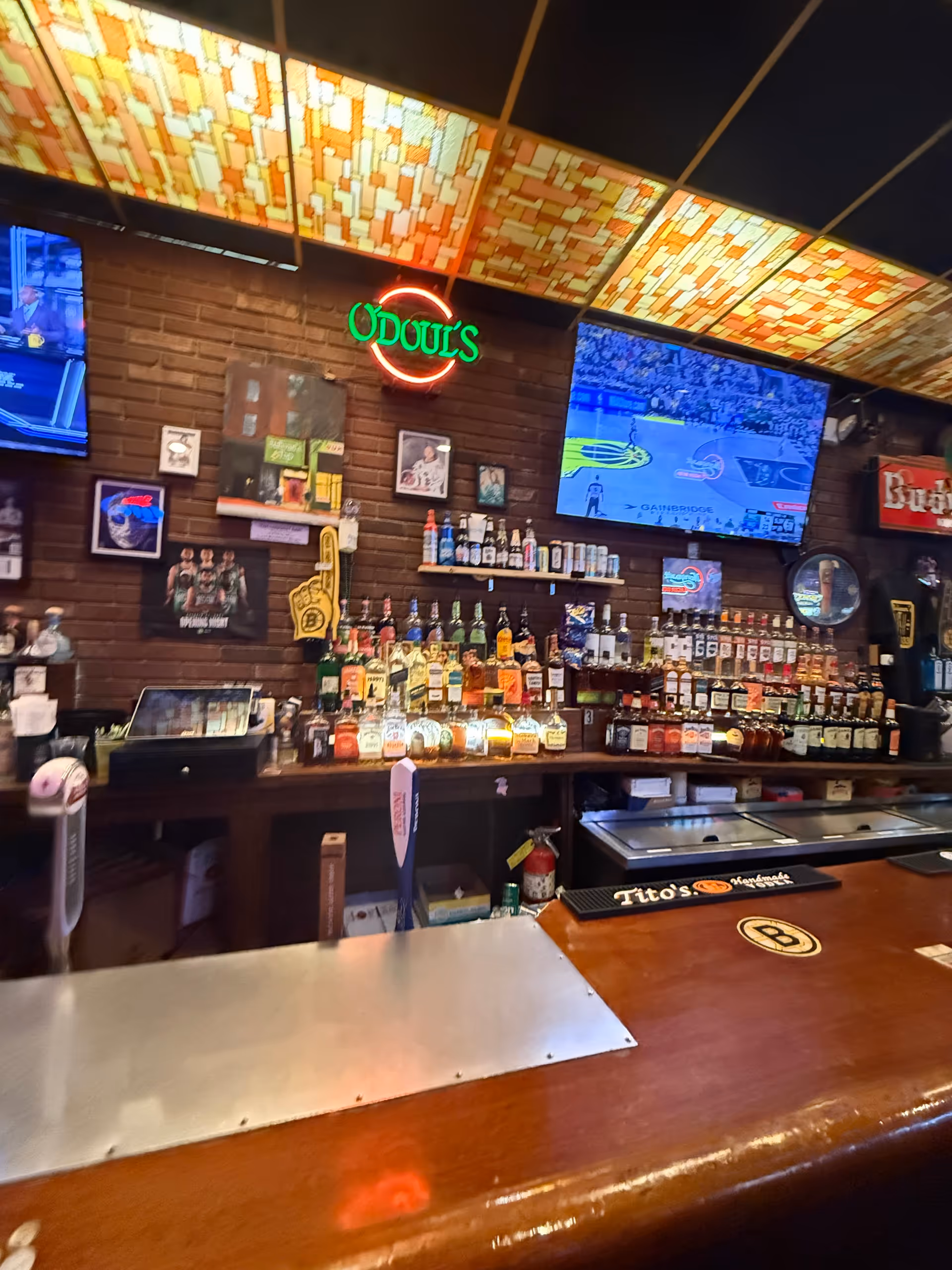 Bar interior with liquor bottles on shelves, brick wall, neon sign, and two TVs.