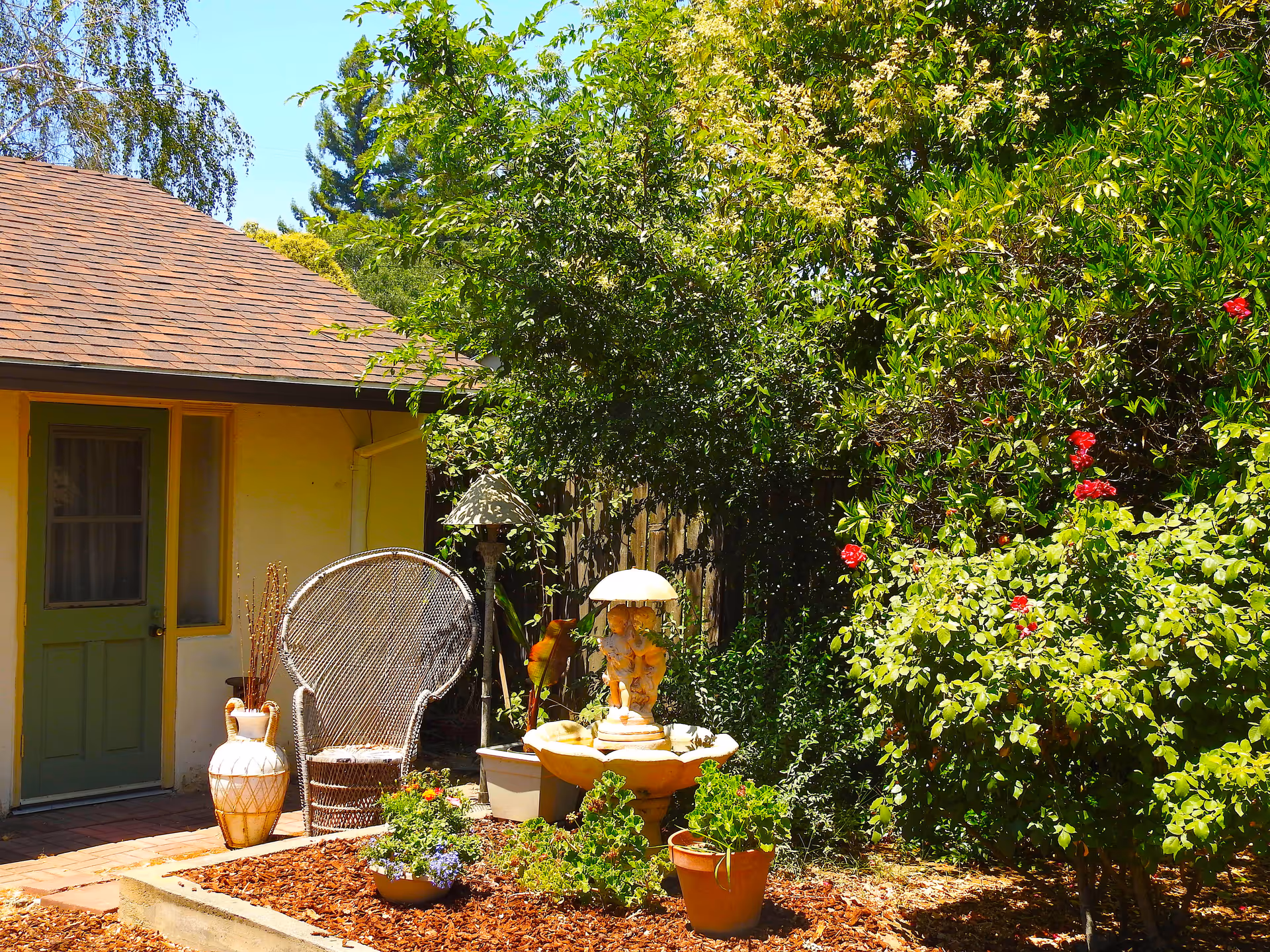 Outdoor courtyard with a yellow house, wicker chair, fountain, and potted plants.