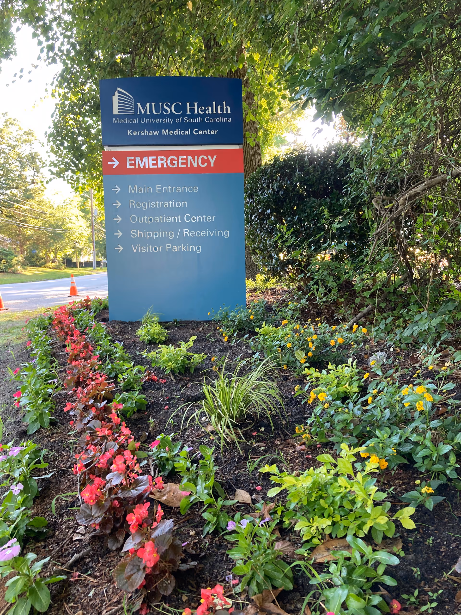 Blue MUSC Health Kershaw Medical Center sign amid blooming flowers and green shrubbery.