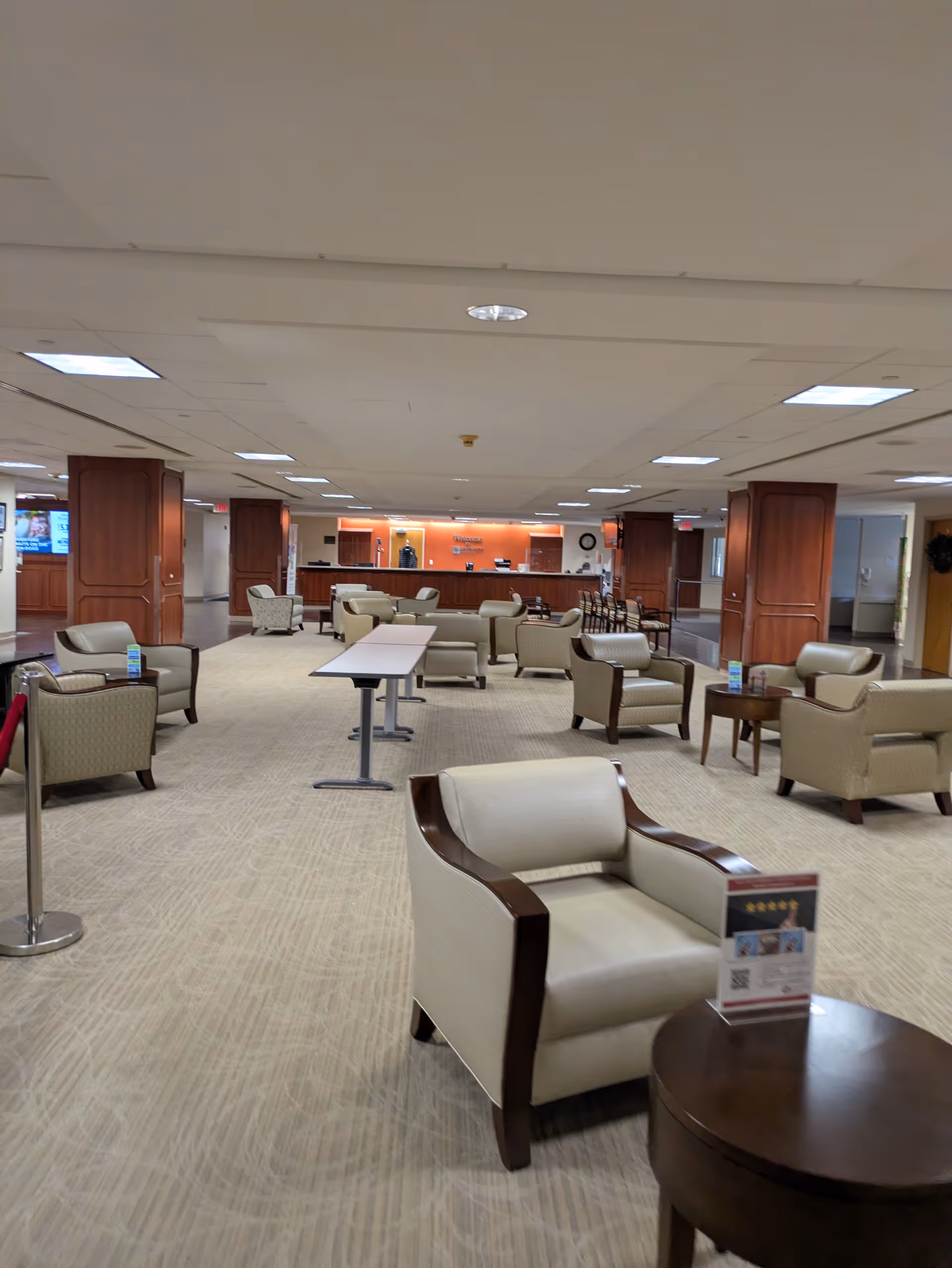 Hospital lobby with beige upholstered chairs, tables, and a distant reception desk.
