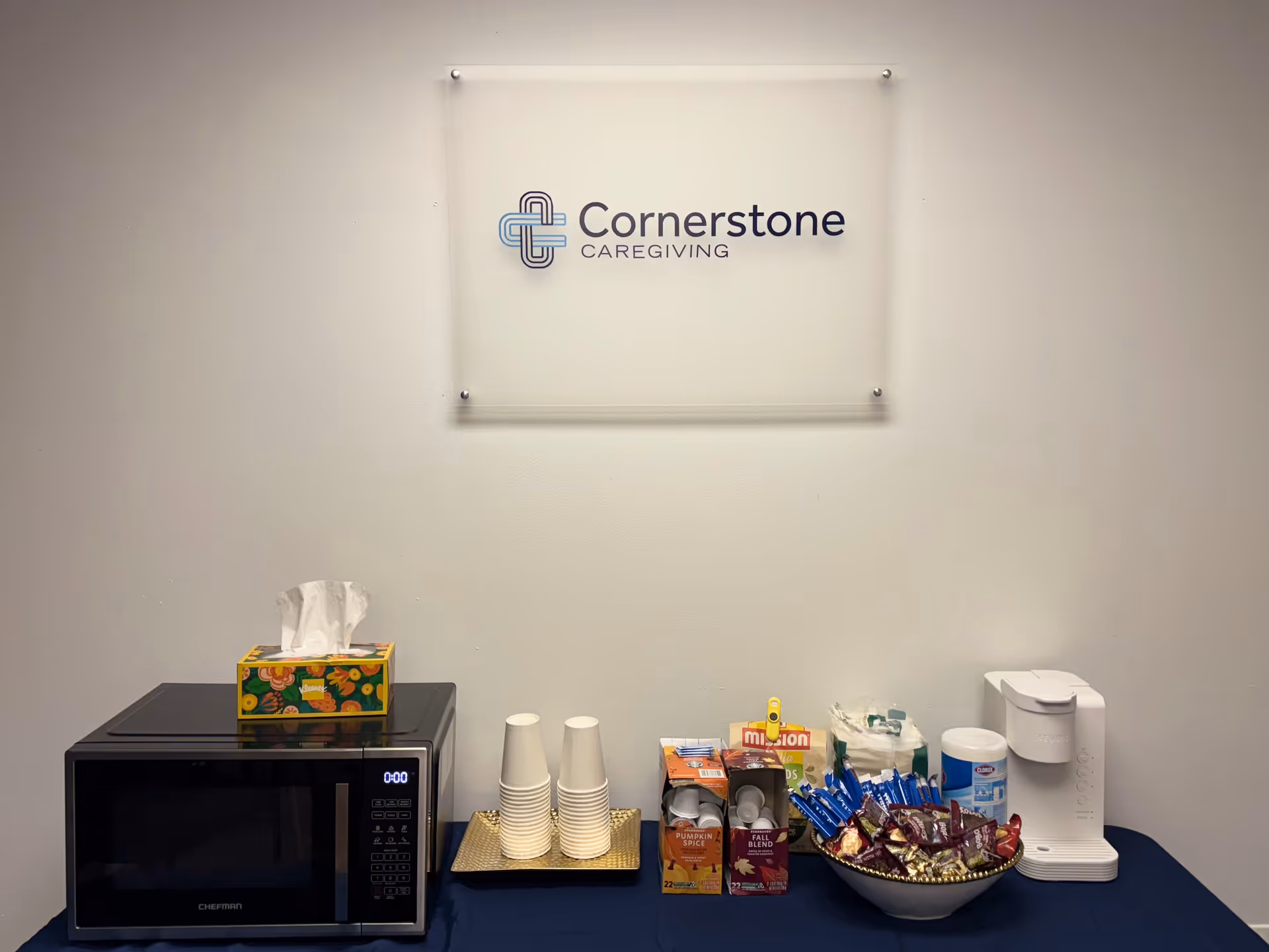 A corner countertop with microwave, cups, snacks, tissue box, and a Cornerstone Caregiving sign.