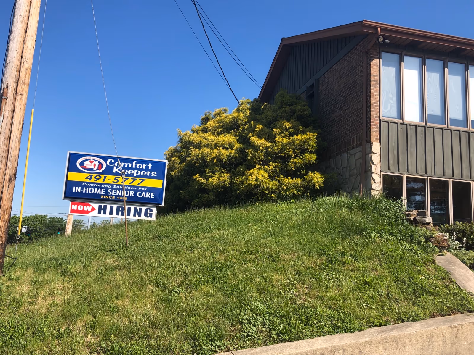 Exterior view of a Comfort Keepers building on a grassy hill with a Now Hiring sign
