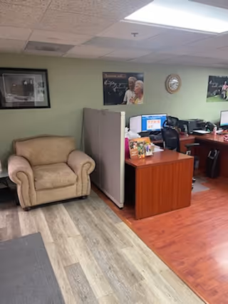 Reception area with beige armchair, partitioned desks, computers, posters, and a wall clock.