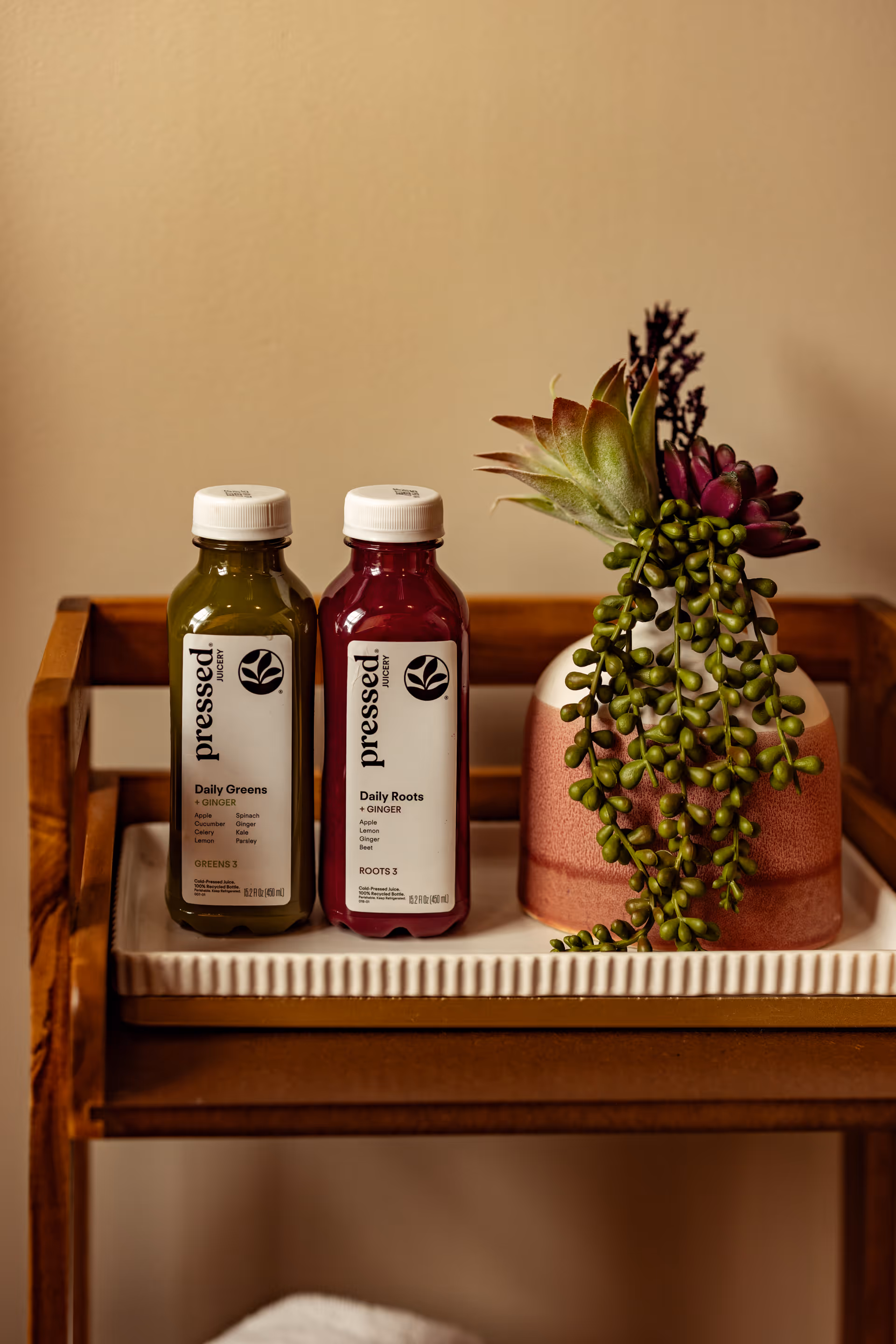 Two juice bottles and a trailing succulent plant on a wooden shelf against a beige wall.