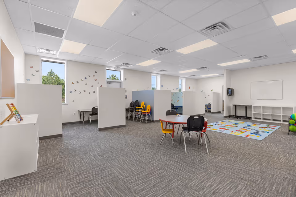 Bright, open ABA therapy classroom with cubicles, small tables, colorful chairs, and a learning rug.