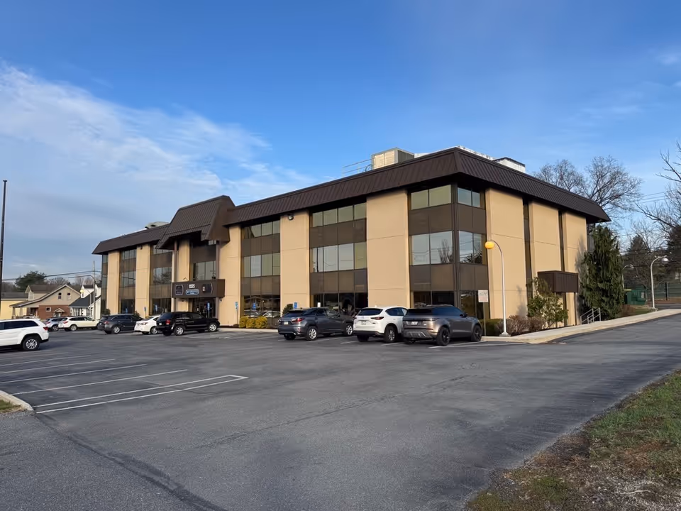 A three-story beige senior living facility with a parking lot under a blue sky.