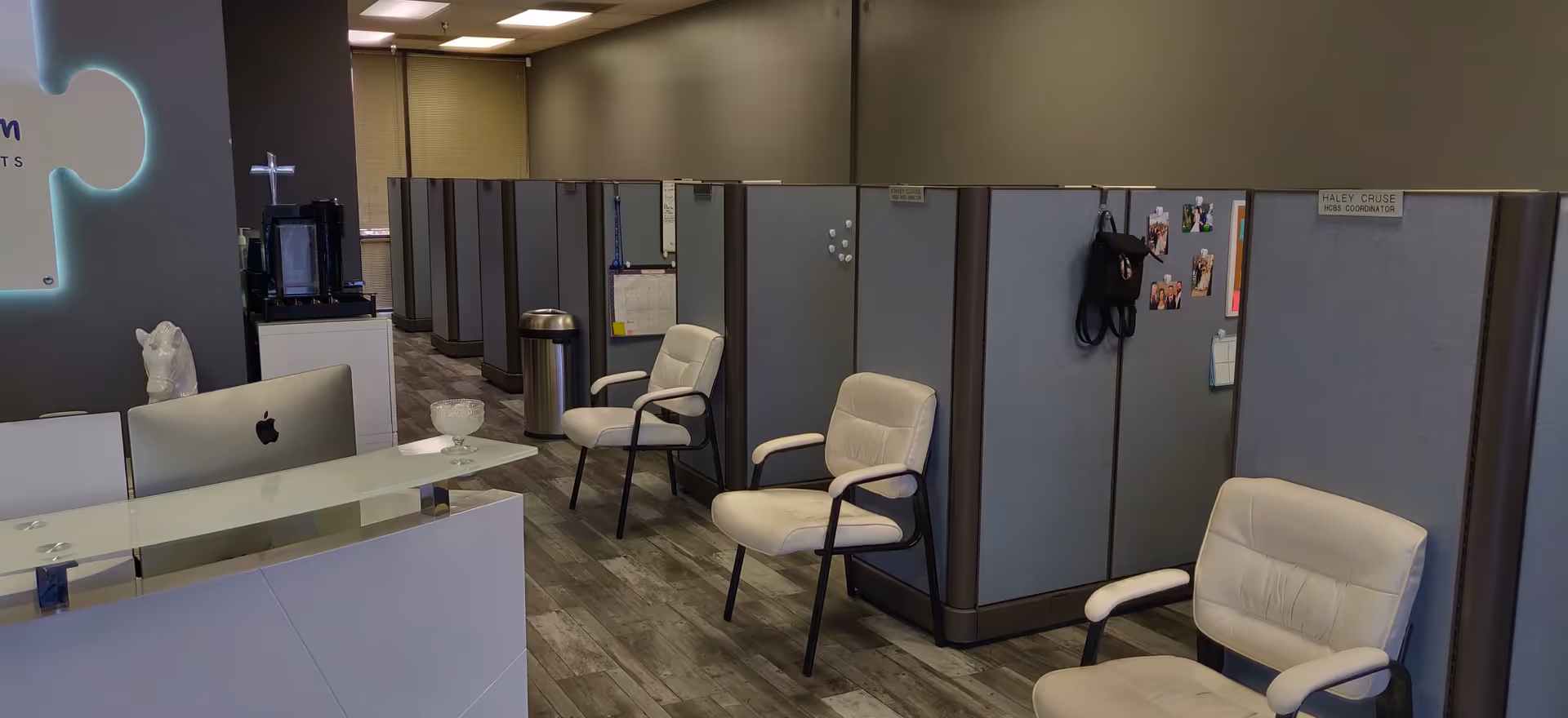Reception area with glass-front desk, white chairs, and office cubicles under gray walls.
