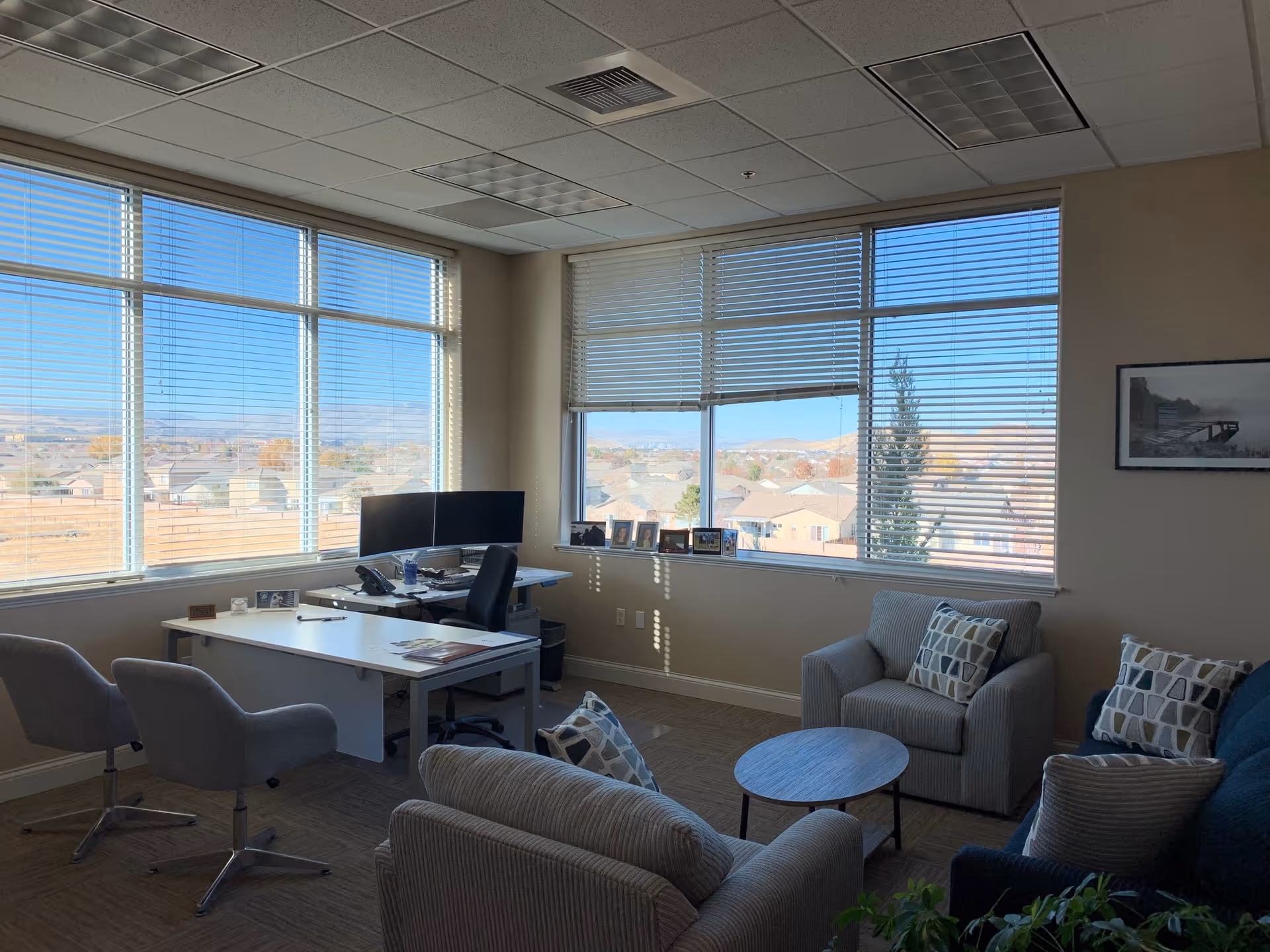 Bright office lounge with a desk, computer, multiple chairs, and large windows overlooking neighborhoods.