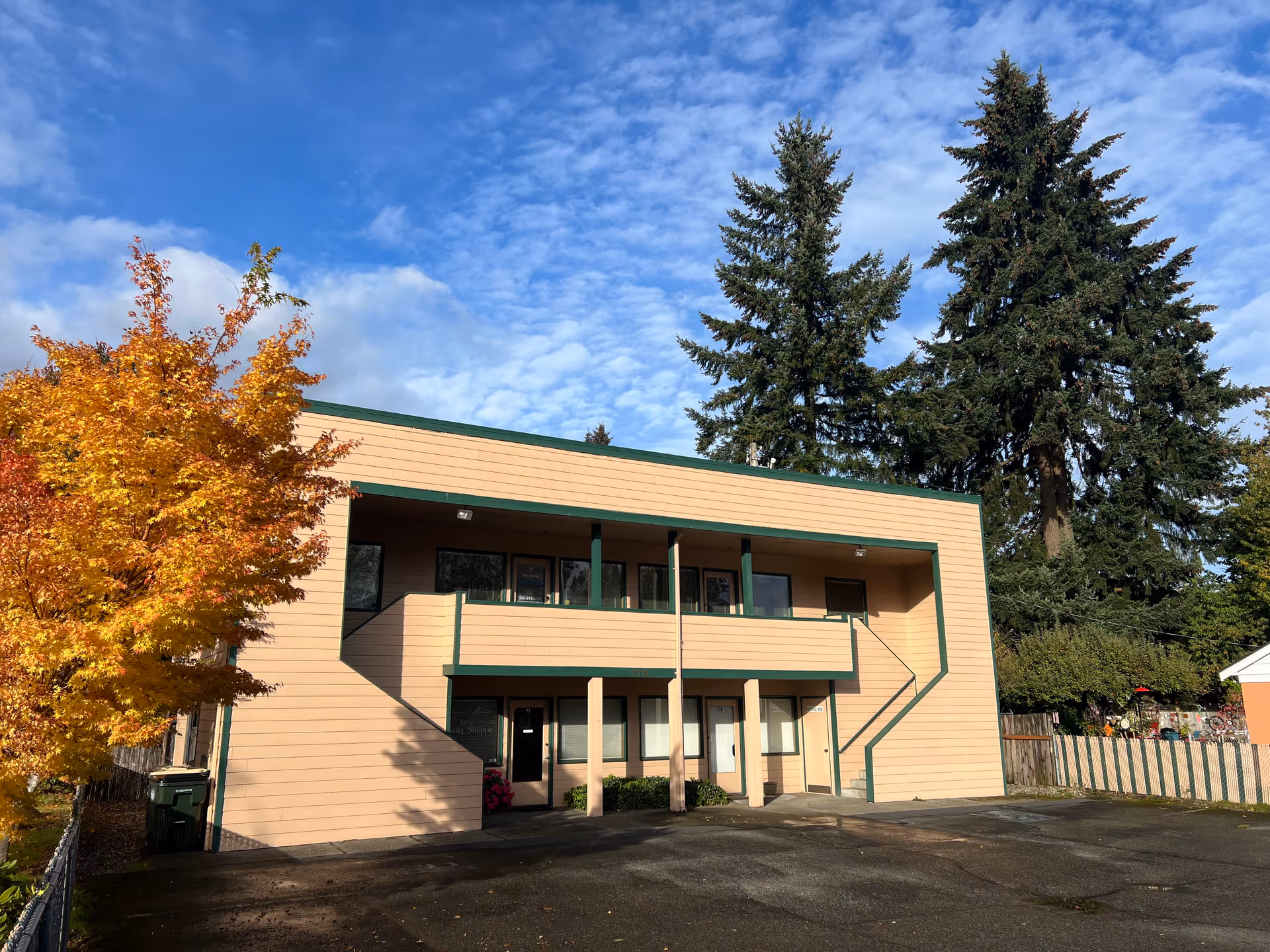 Two-story beige building with green trim, stairs, parking area, and autumn trees under blue sky.