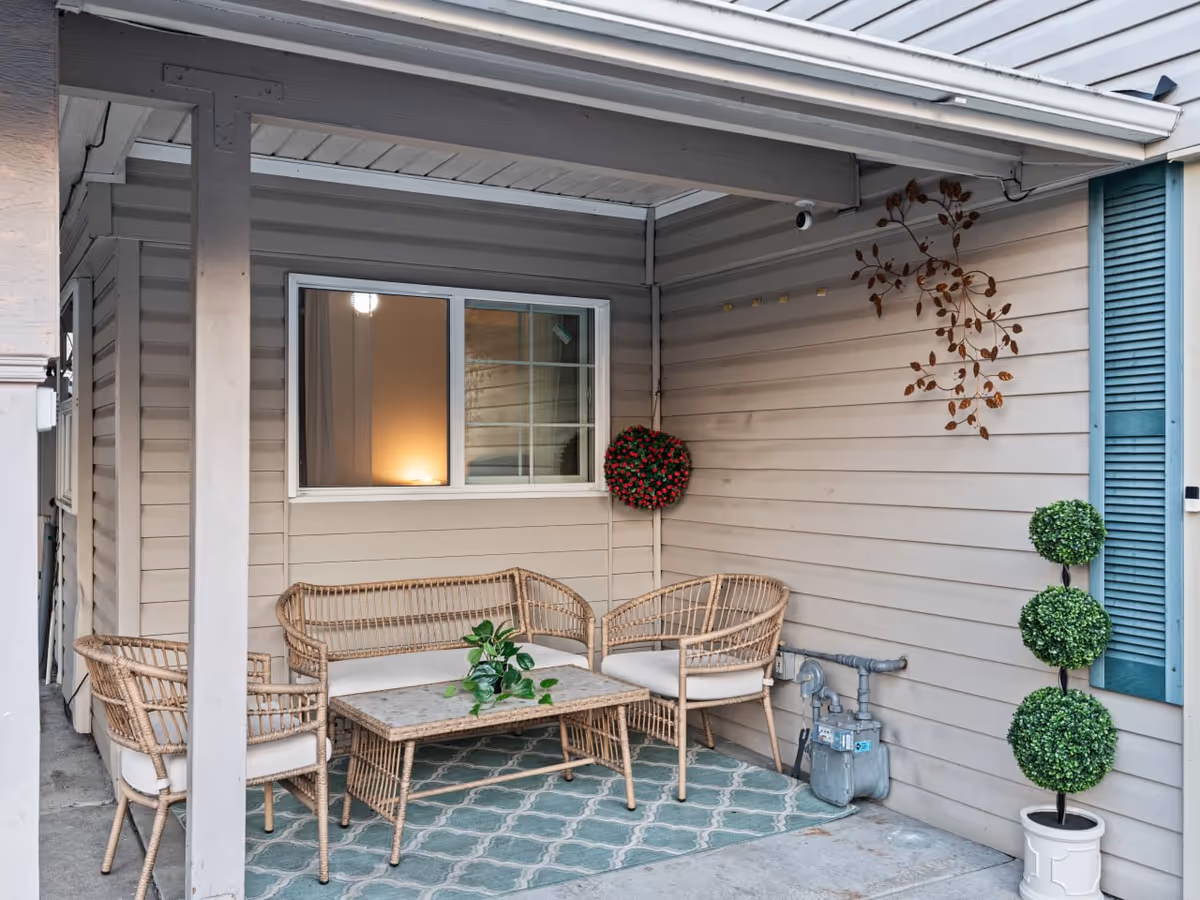 Covered outdoor patio with wicker chairs and a table beside a beige house.