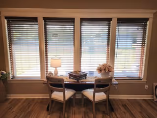 A cozy dining nook with a small table, two chairs, lamp, and large windows with blinds.