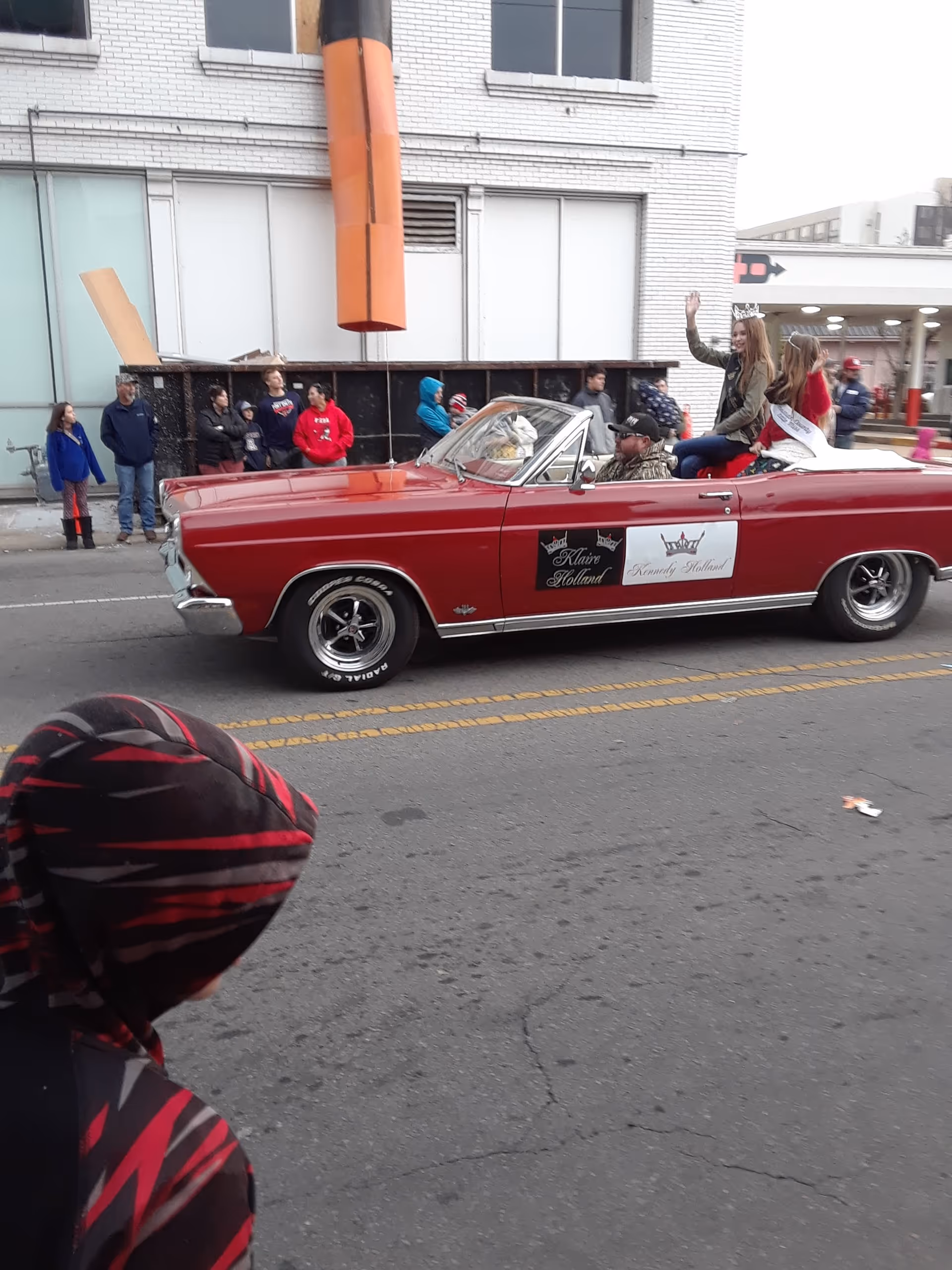 Red vintage convertible with riders drives through a crowded outdoor street parade.