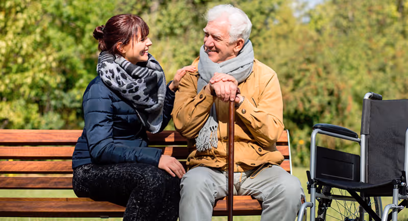An elderly man on a bench outdoors chats with a smiling caregiver, wheelchair beside them.