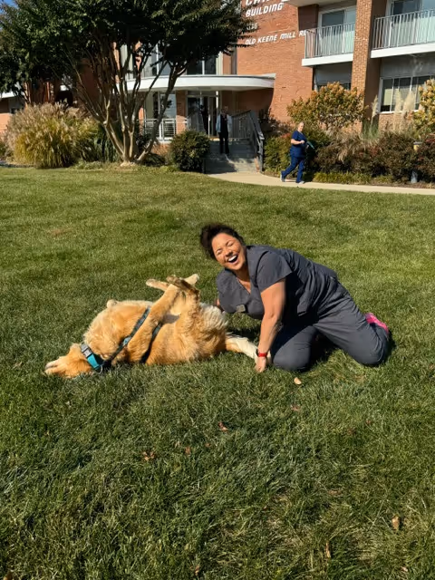 Smiling caregiver plays with a dog on the grassy lawn outside Visiting Angels Senior Home Care.