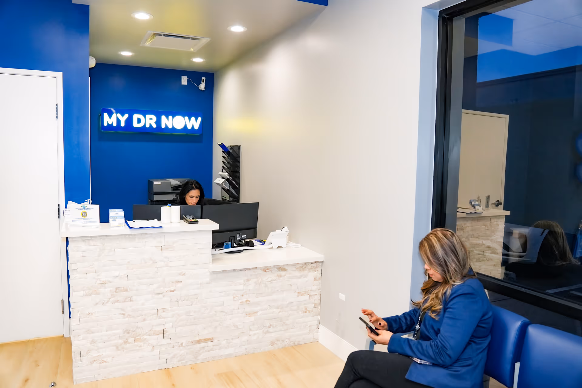 Reception area of My Dr Now with blue wall, sign, and a seated visitor.