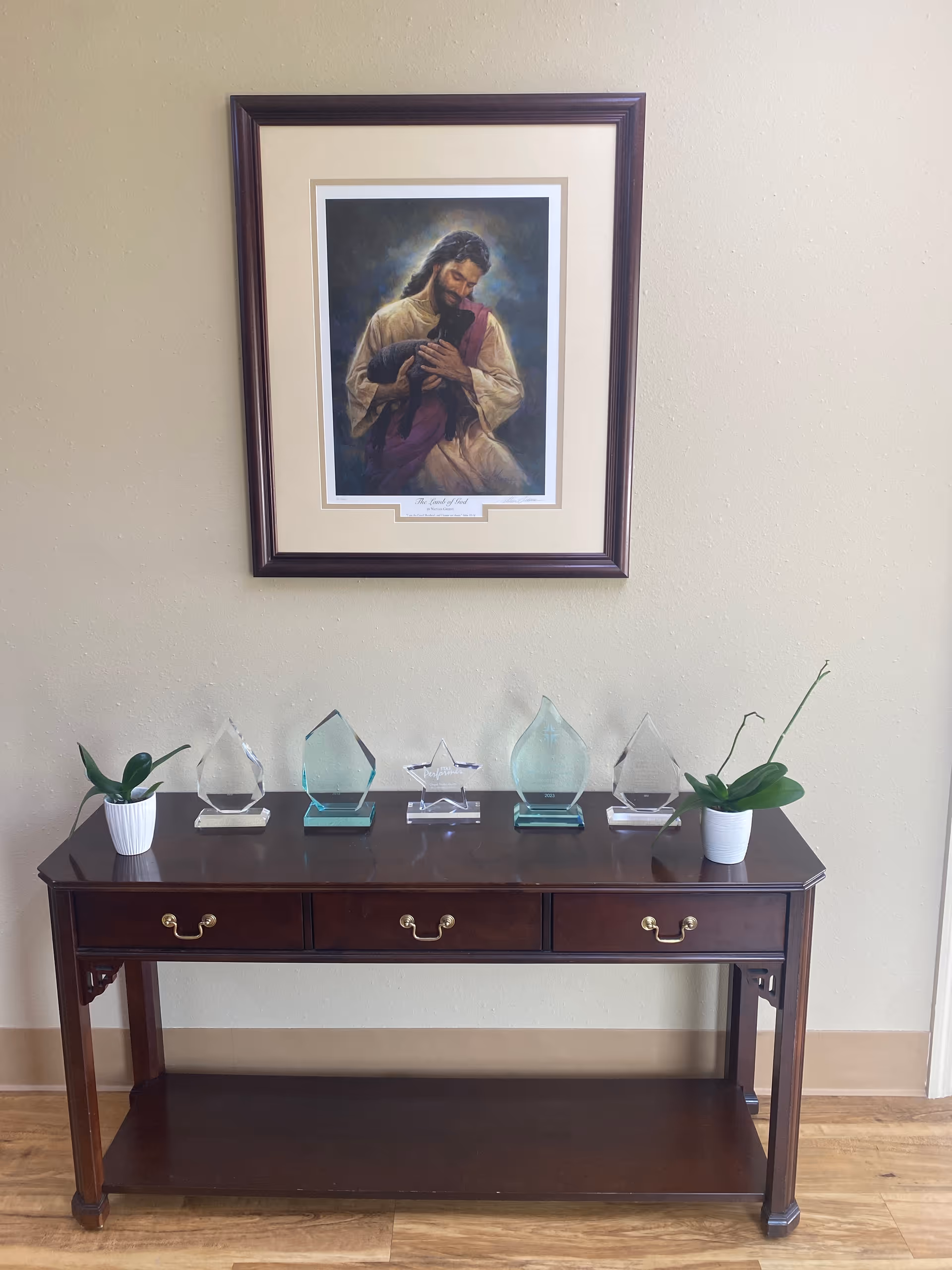 Wooden console table against beige wall with framed religious portrait above and glass awards.