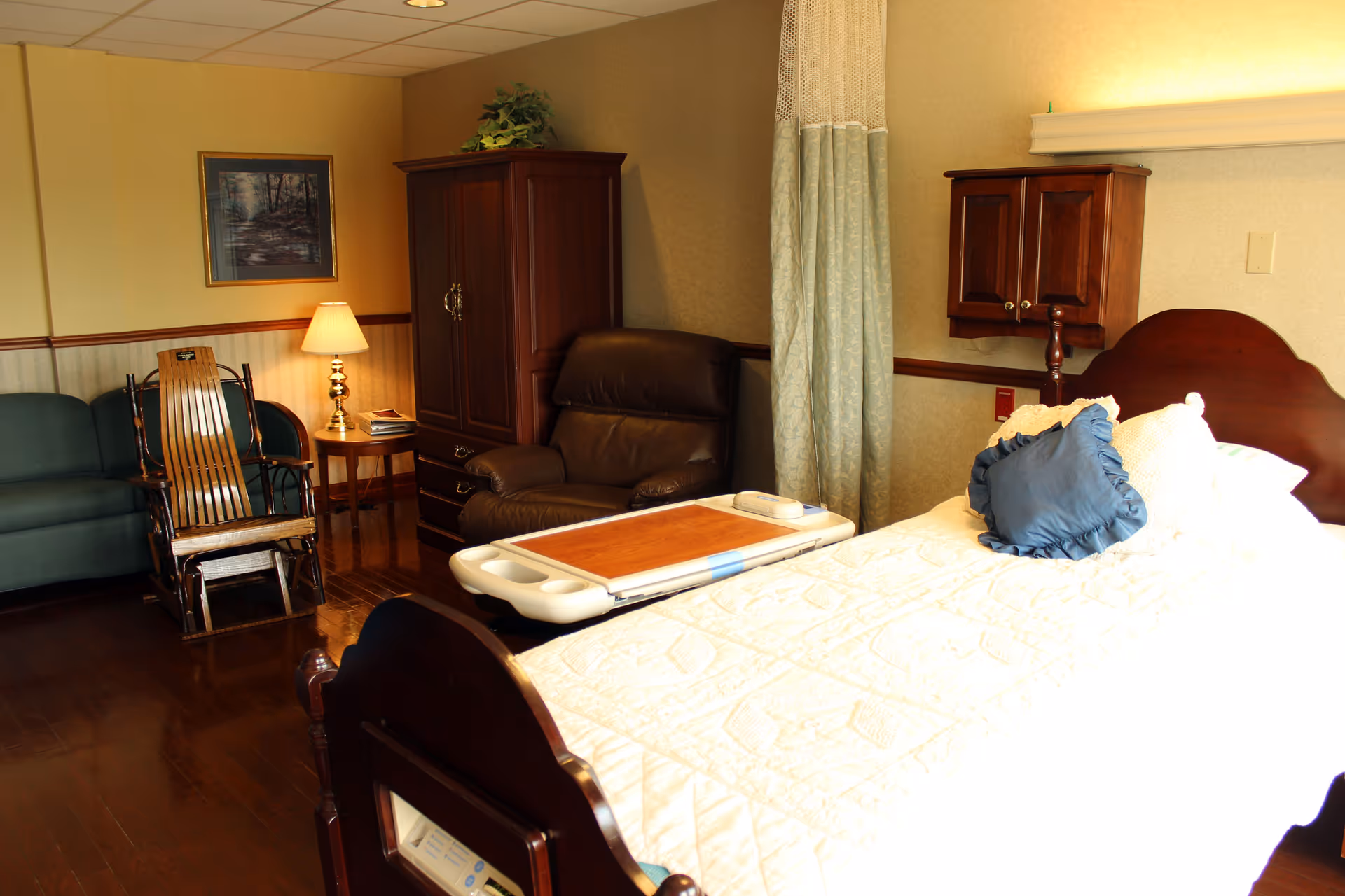 Bedroom in a senior facility featuring a bed, seating area, wardrobe, and privacy curtain.