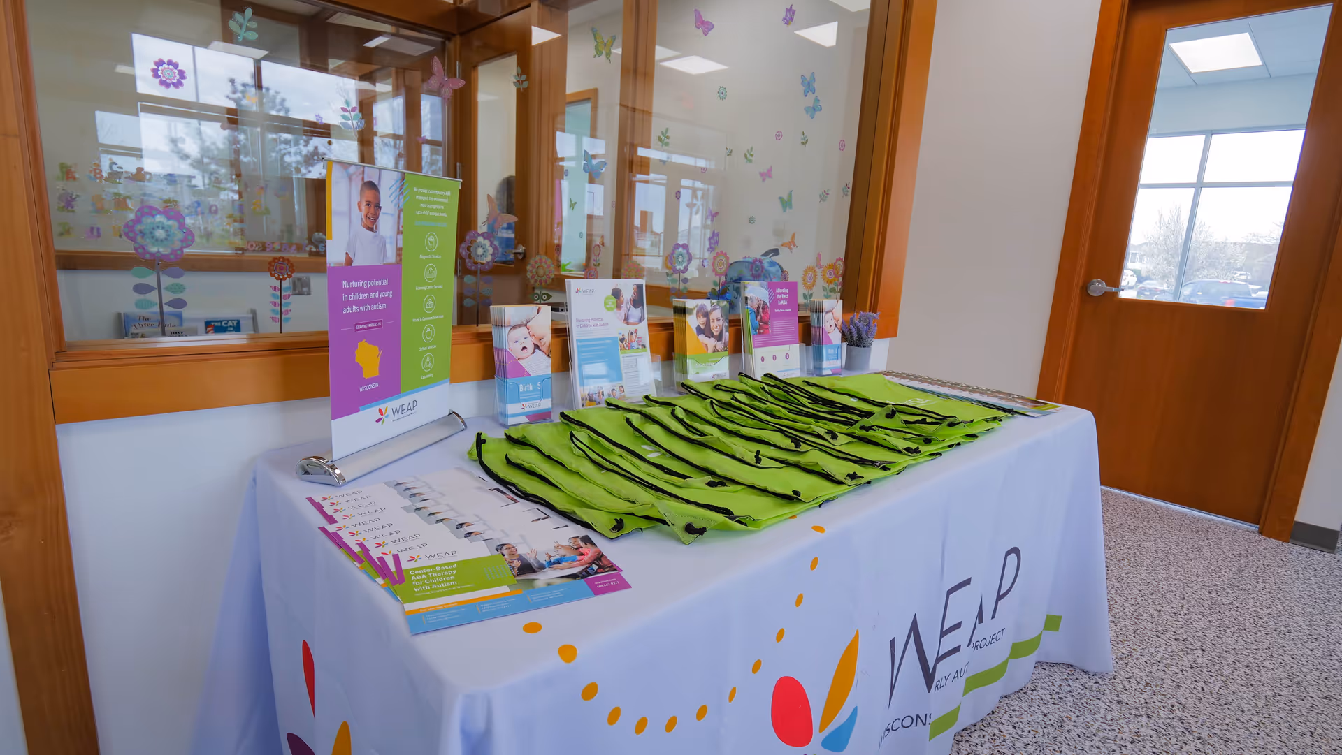 Promotional display table with WEAP materials and green tote bags in a facility lobby.