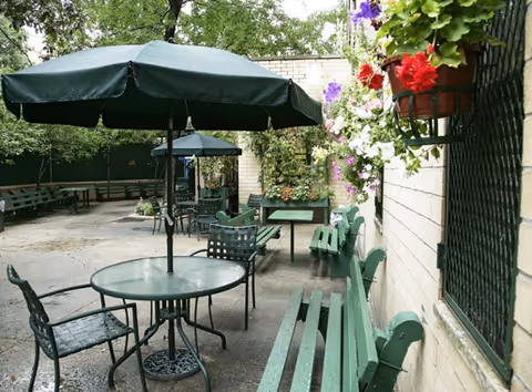 Outdoor patio with metal tables and chairs under umbrellas beside a brick wall and flowers.