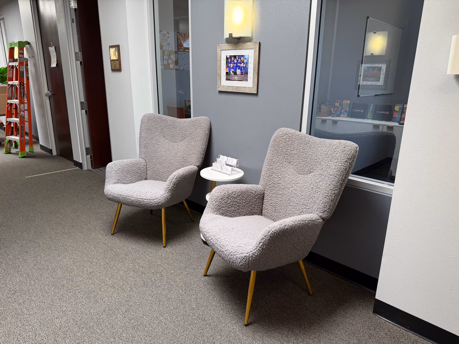 Two grey upholstered chairs with a small round table in a hallway seating area.