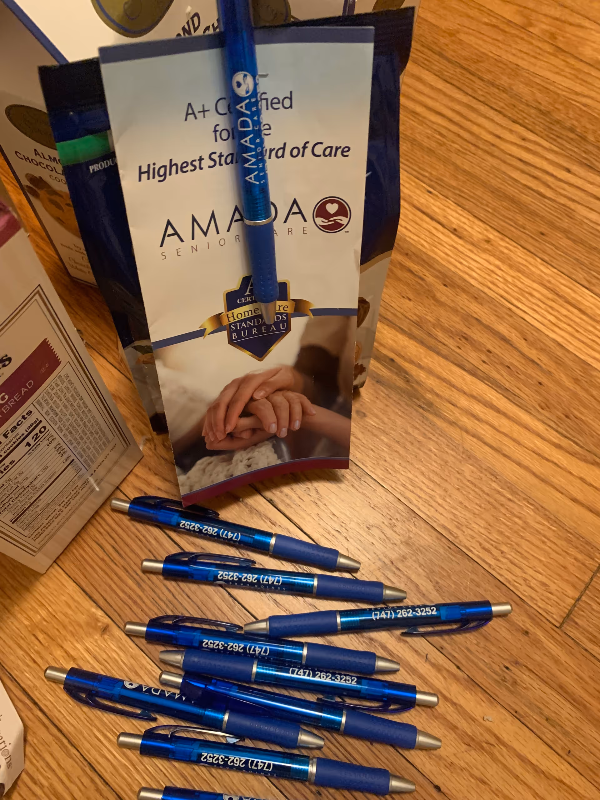 Blue promotional pens and Amada Senior Care brochure scattered on a wooden floor