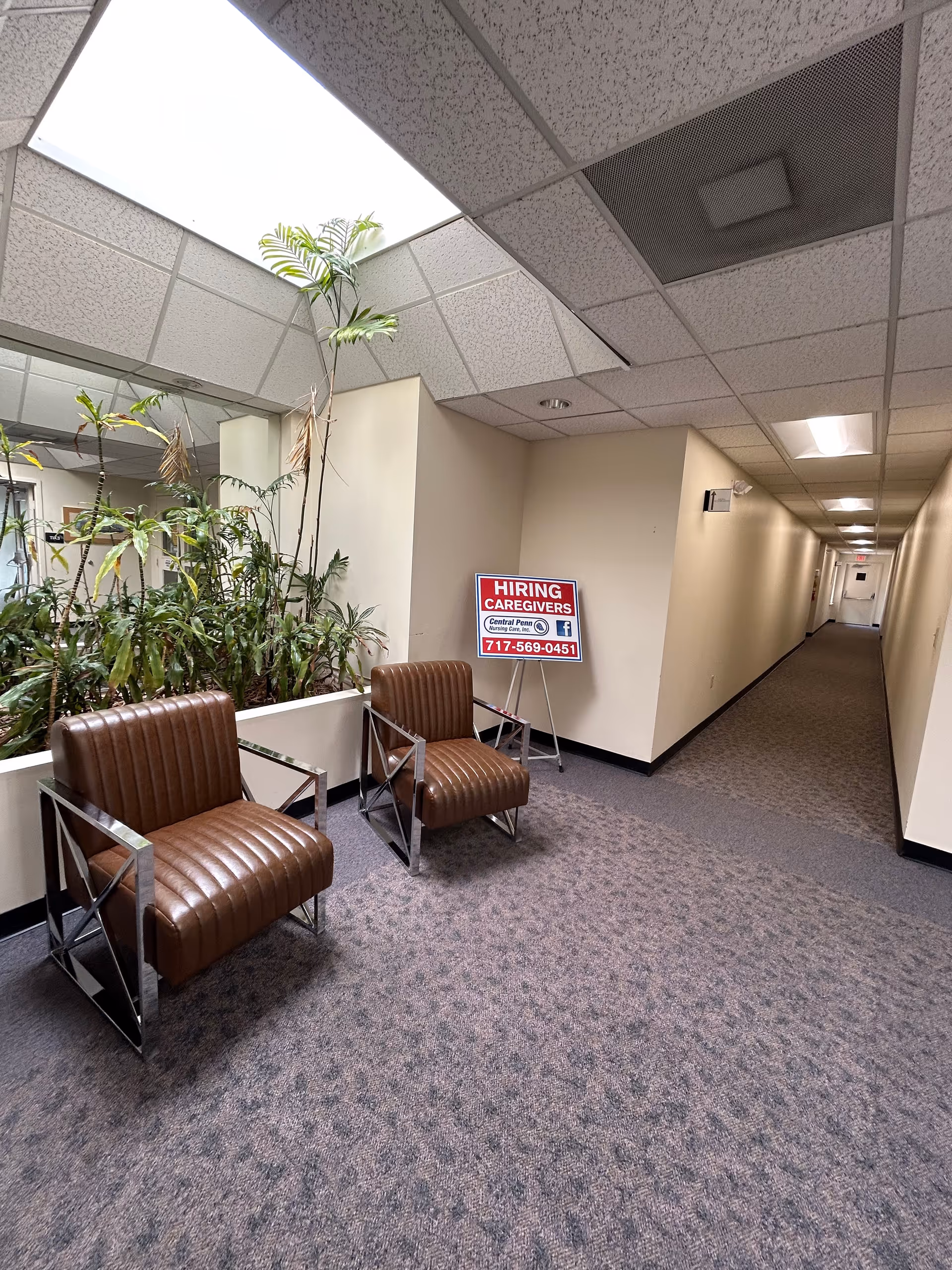 Indoor nursing facility hallway with two brown chairs, plants, and a hiring sign.