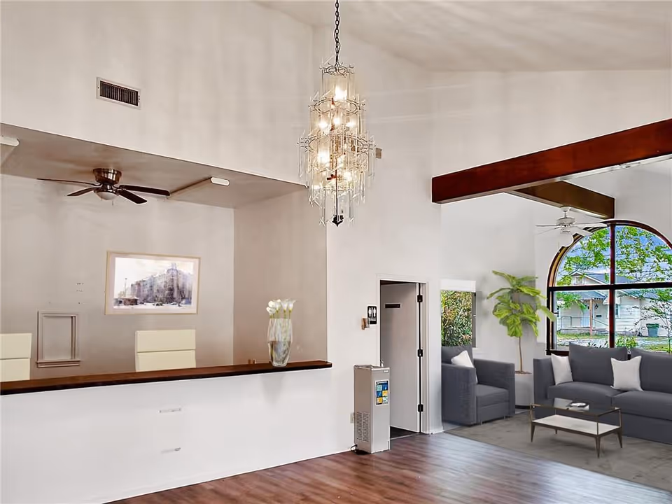 Reception area with a chandelier, high ceiling, wooden floor, and gray seating by an arched window.