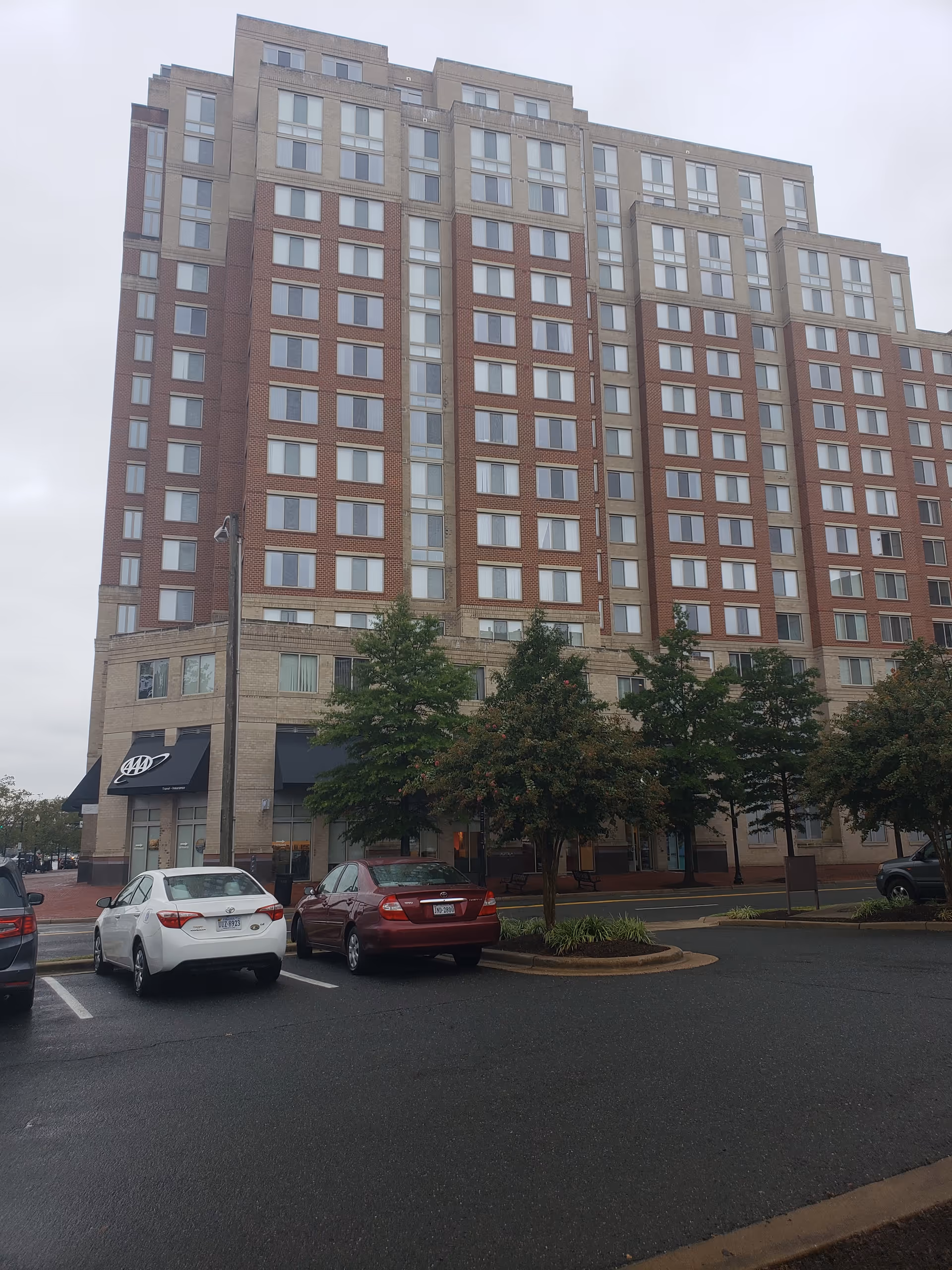 Tall brick senior living building with parked cars and trees along the entrance.