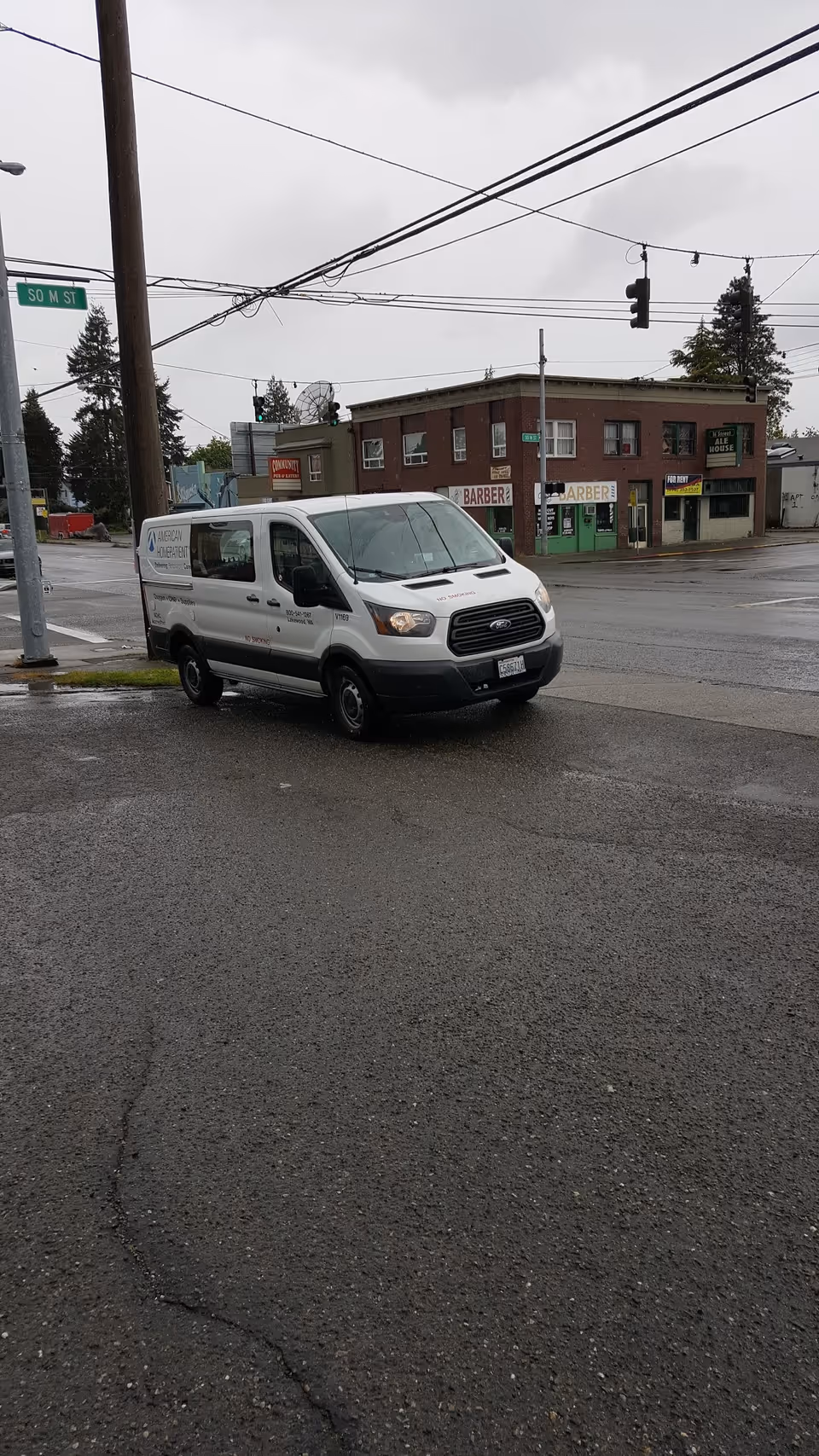 White van parked on a wet urban street with brick storefronts and barber shops in background.