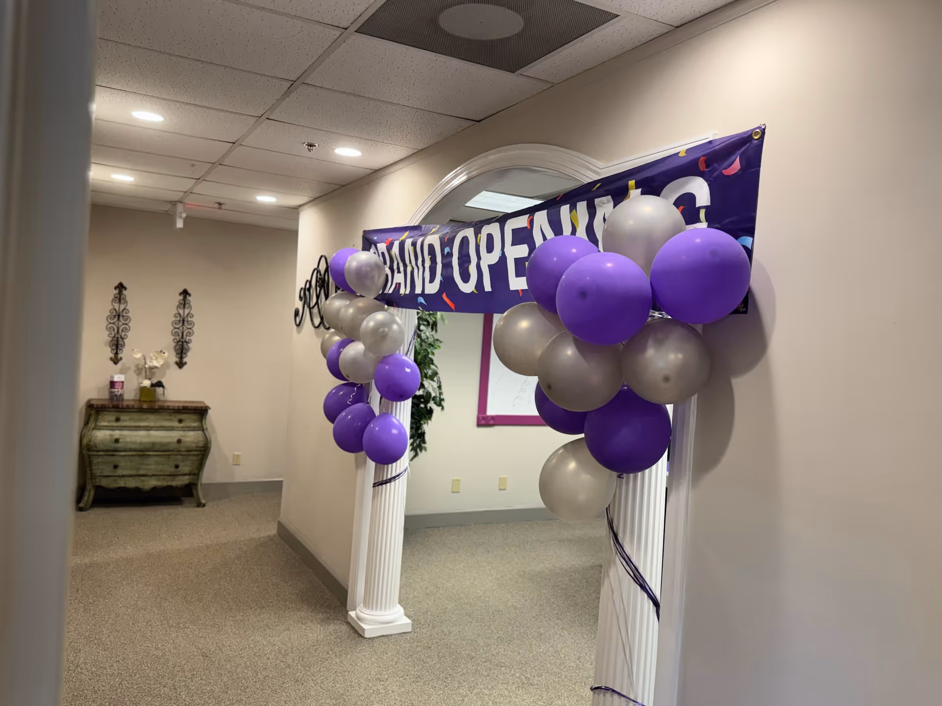 Interior hallway with purple and silver balloons framing an arch and grand opening banner.
