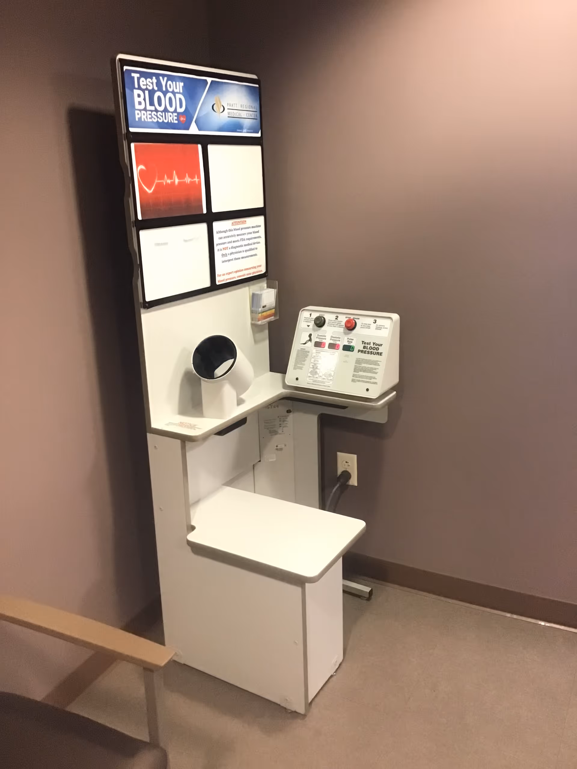 Blood pressure testing station with monitor in a beige hallway.